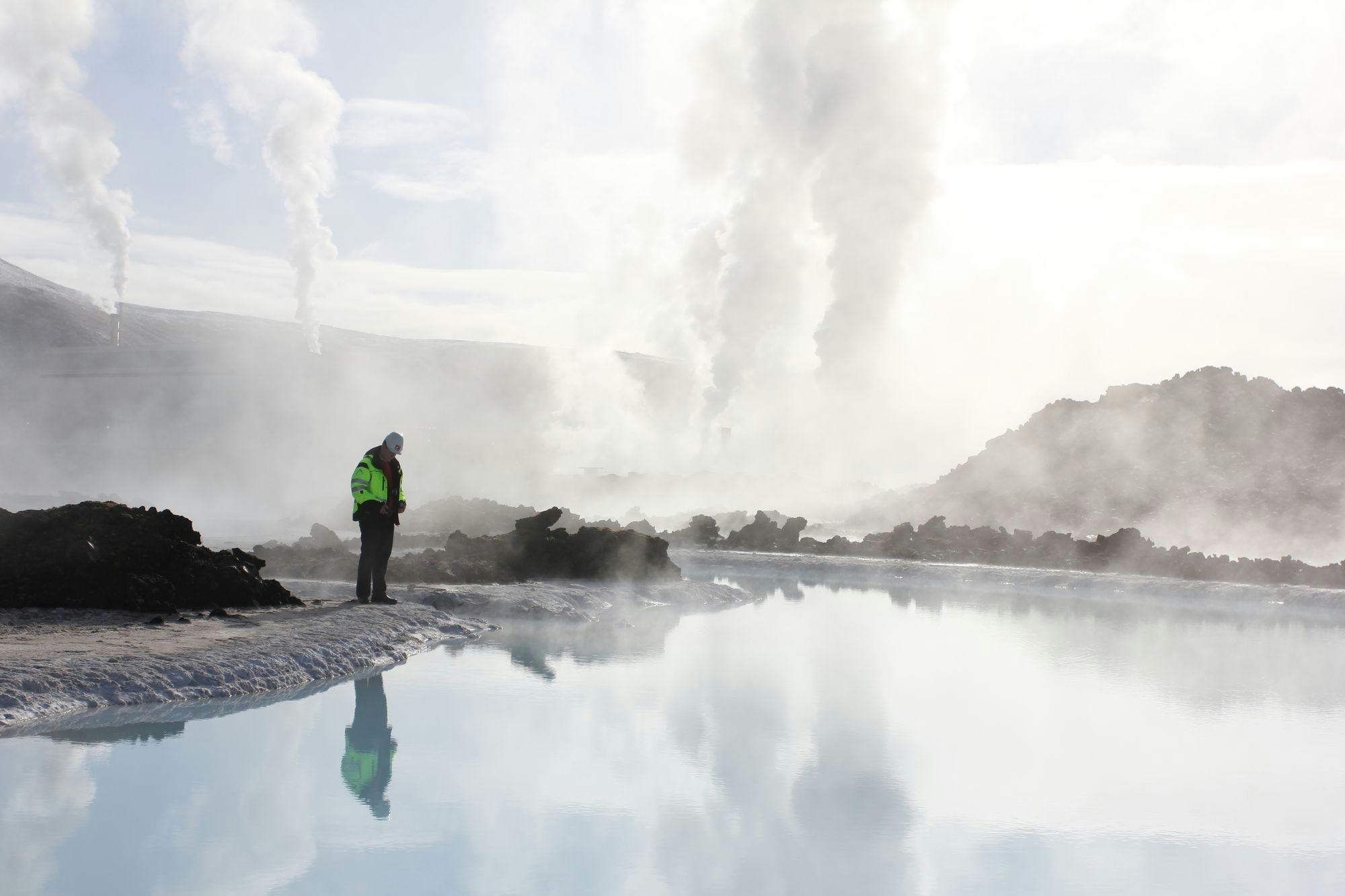 A man in workwear stands by hot water in a rough terrain, hot springs in the background