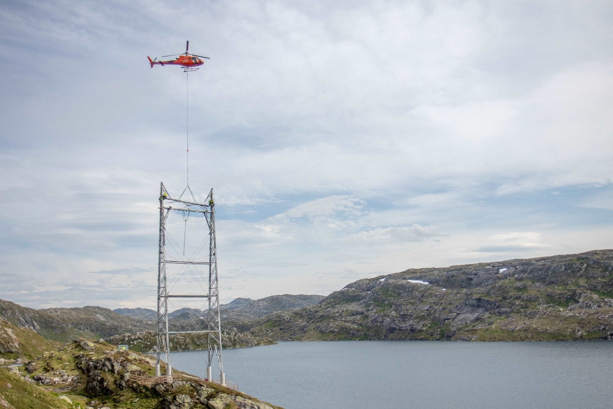 A mast that is being erected hangs from a helicopter in beautiful nature
