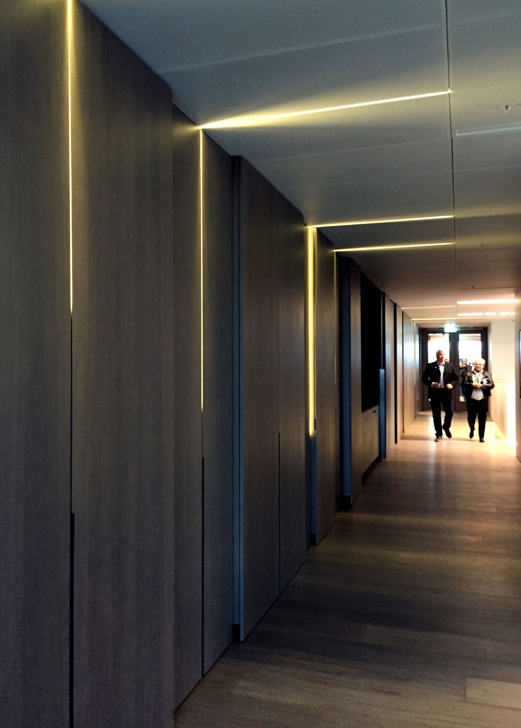 Two people facing a hallway with wood-colored walls with lit stripes going up into the ceiling