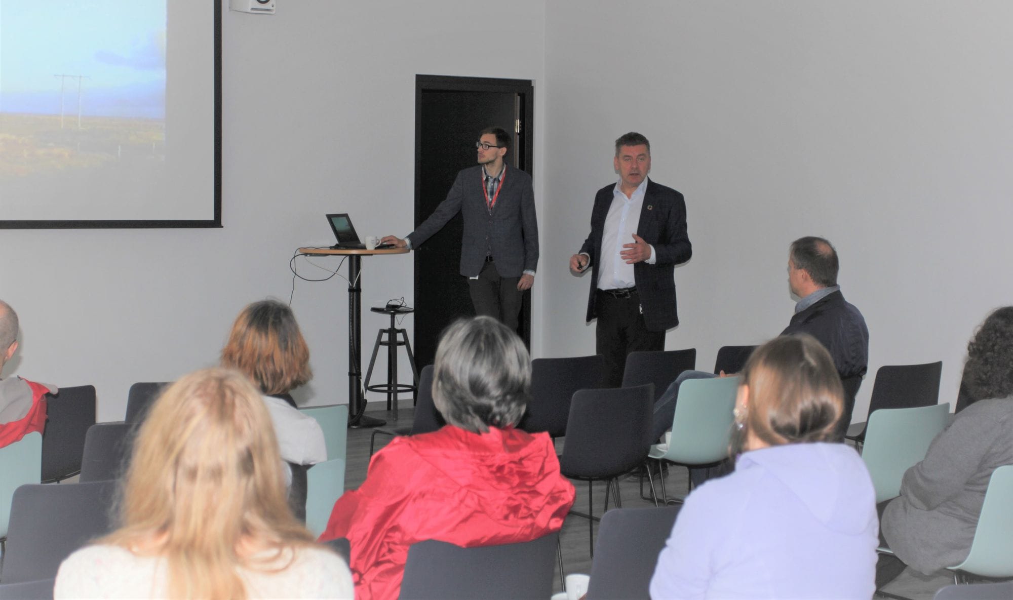 A man giving presentation to a group of people and another man staring to a laptop behind him