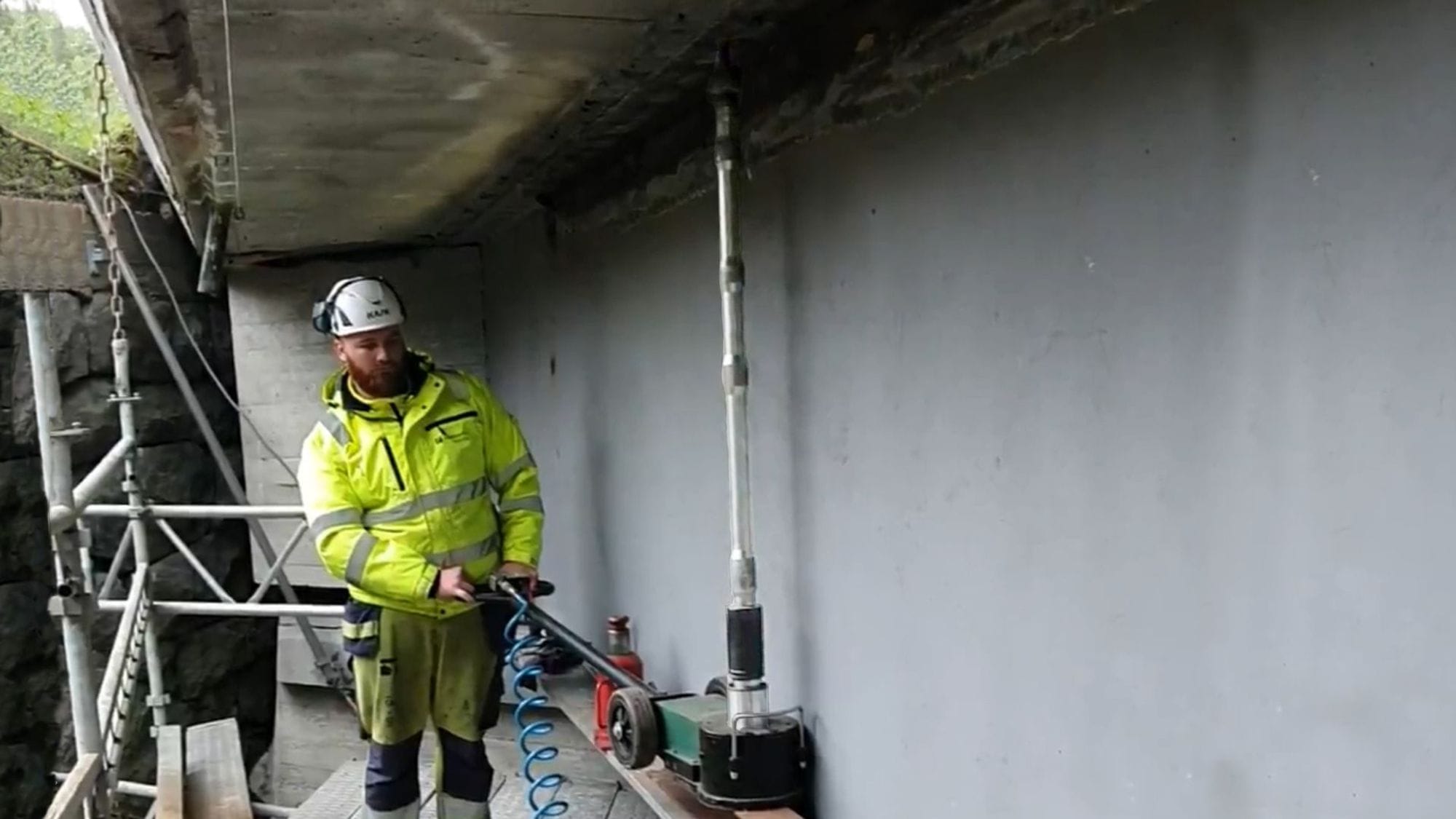 A worker in yellow work clothes with a white safety helmet working with equipment under a bridge.