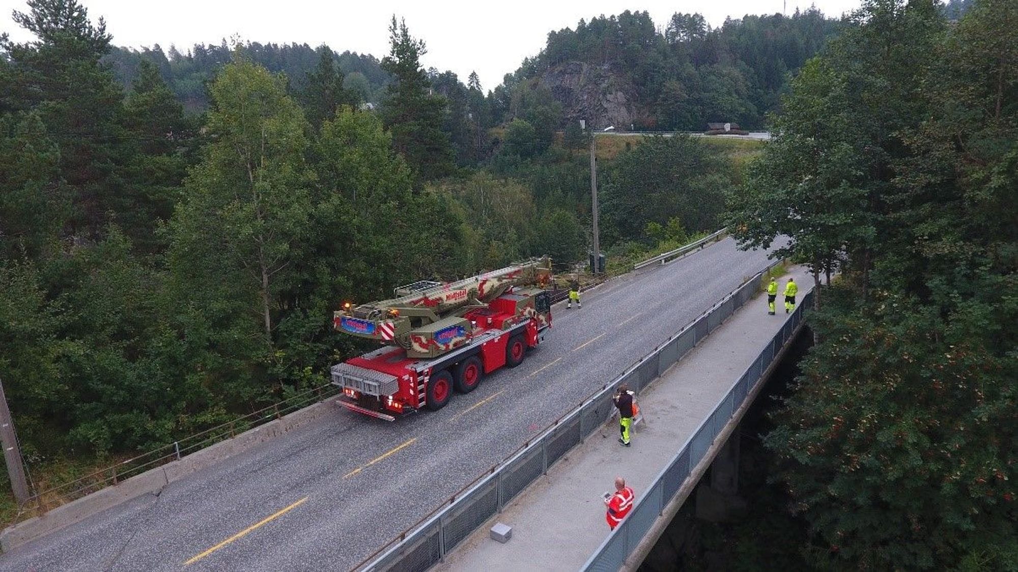 A concrete bridge surrounded by spruce trees. A red basket truck is standing on the bridge and four men in yellow and red work clothes.