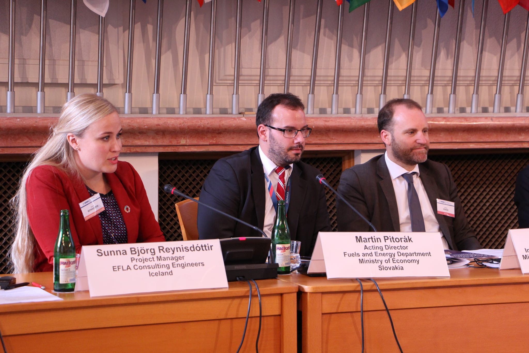 A lady in red blazer speaking in a meeting, seated next to two men in black blazer
