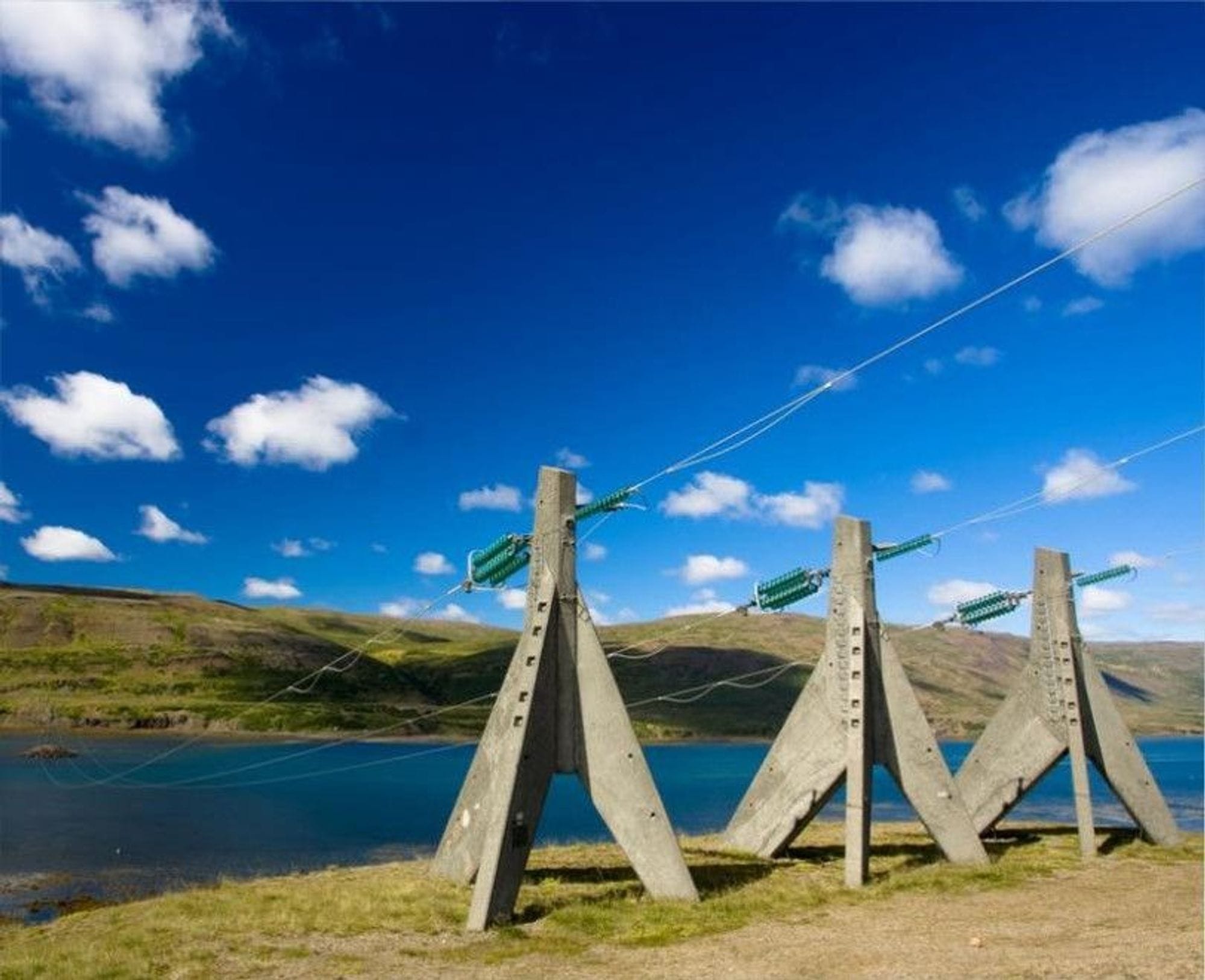 Three concrete electric towers side by side in front of water and green slopes