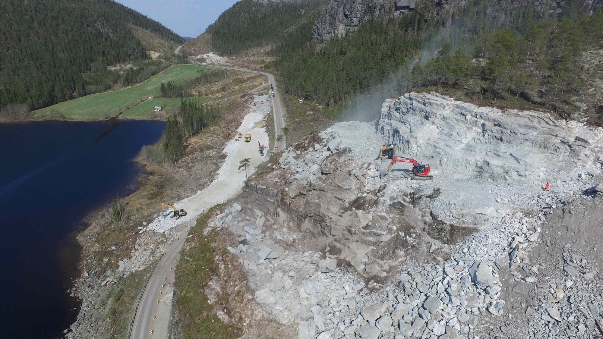 Two excavators working in rough and light grey rocky terrain and road and moutains in the background