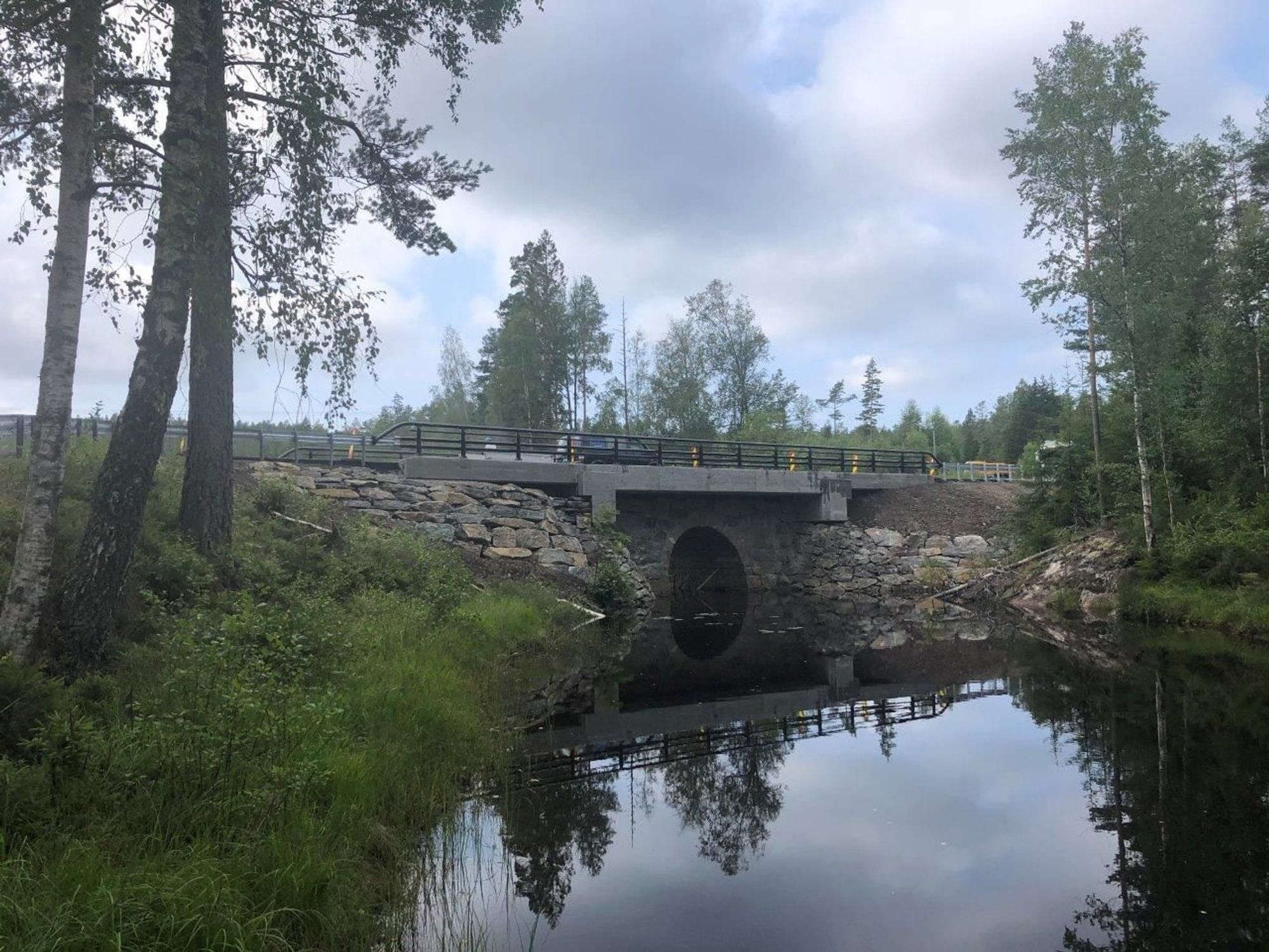 Renewed Sunds bridge. A bridge that is partly stone-paved and partly concrete on the top. Still water passes through the bridge's circular openings and trees and partly cloudy skies around the bridge.