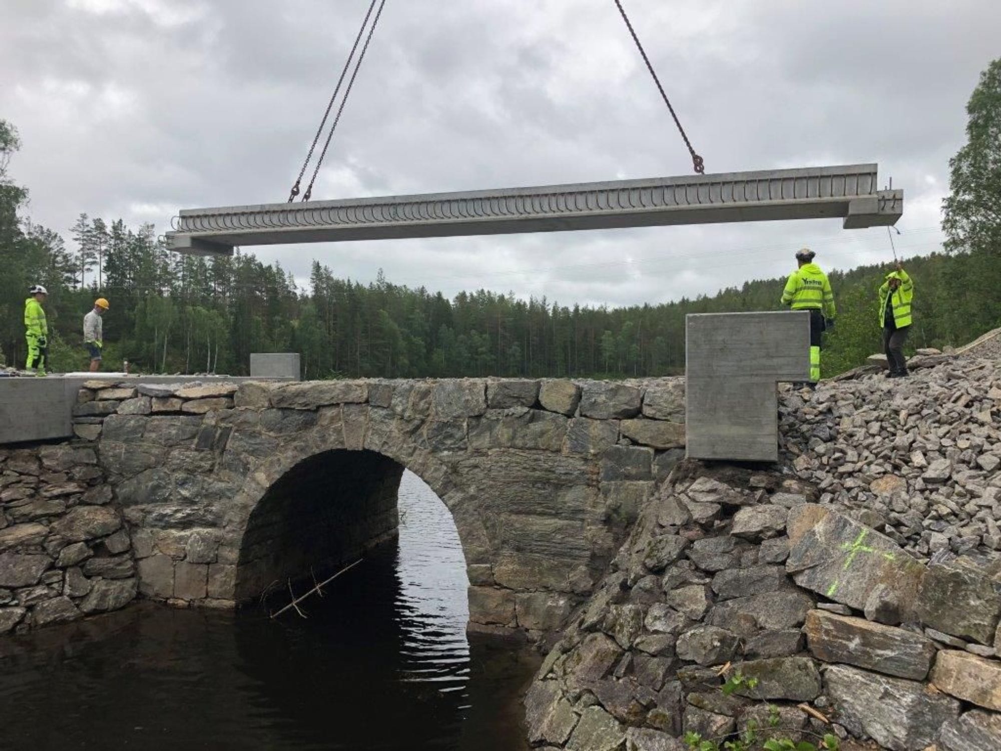 Four men standing on top of nearly finished Sunds bru built partly of stones.  Concrete beam is hanging from vires and being installed for the new superstructure. 
