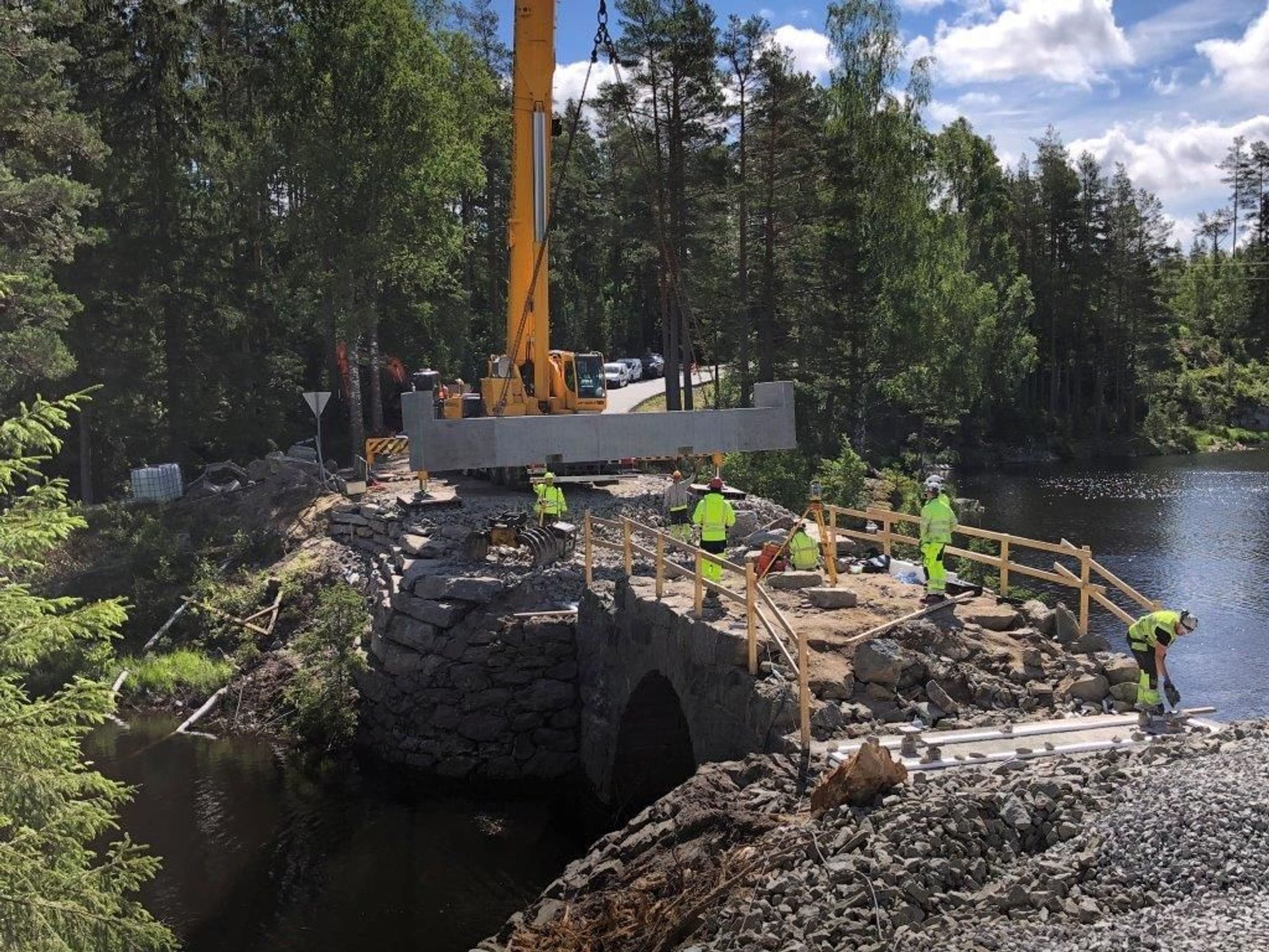 Orange crane works on the installation of new land tanks  on the bridge. Men in yellow work clothes are on top of the bridge at work.