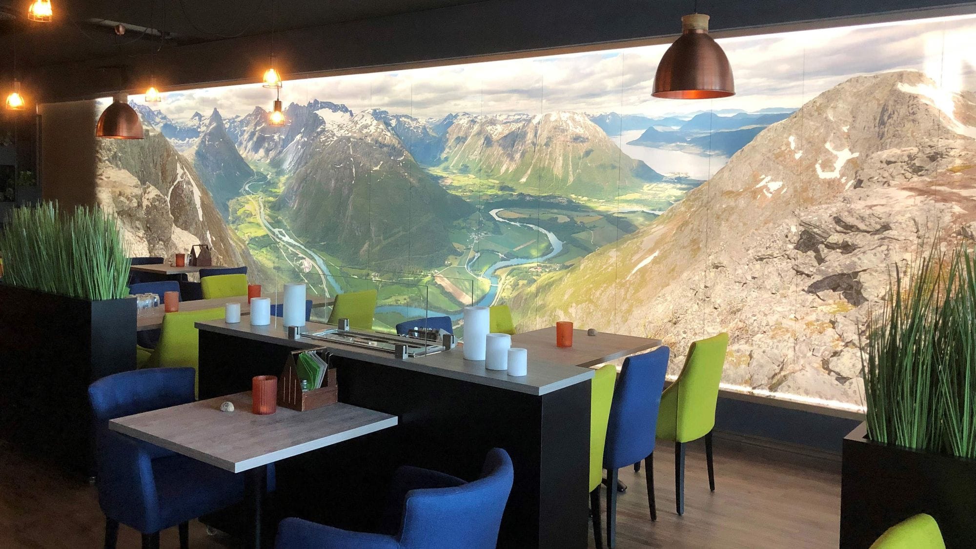 Tables and blue and green chairs in a dark space and a big picture wall with lighting that shows magnificent mountain surroundings