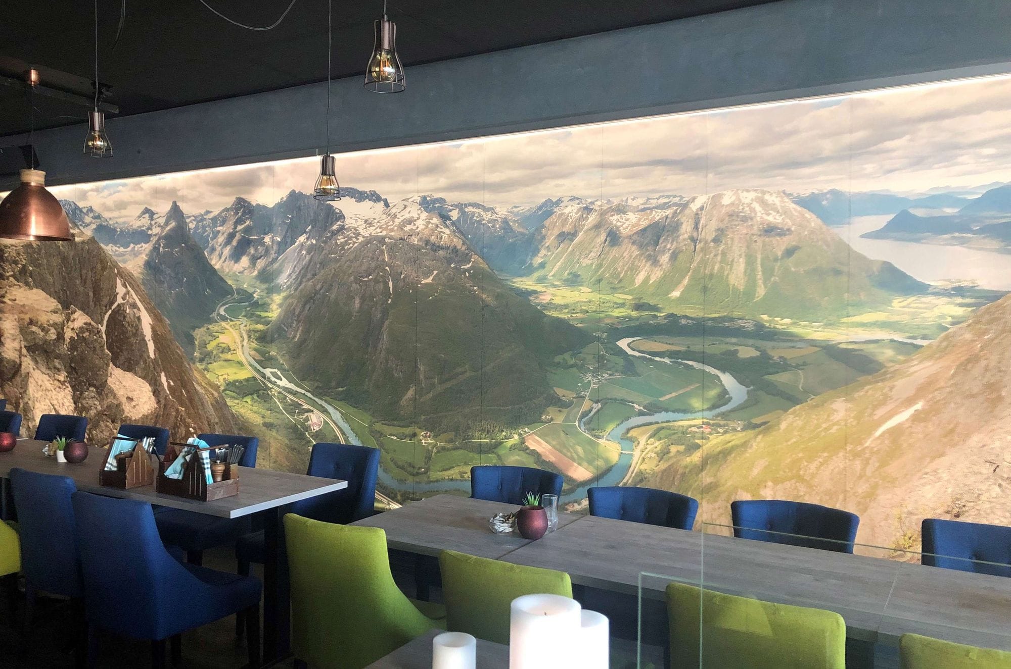 Long tables with blue and green chairs in a dark space with a long wall piece on the wall that is a picture of mountains and nature and has a lighting in the picture that makes the picture pop.