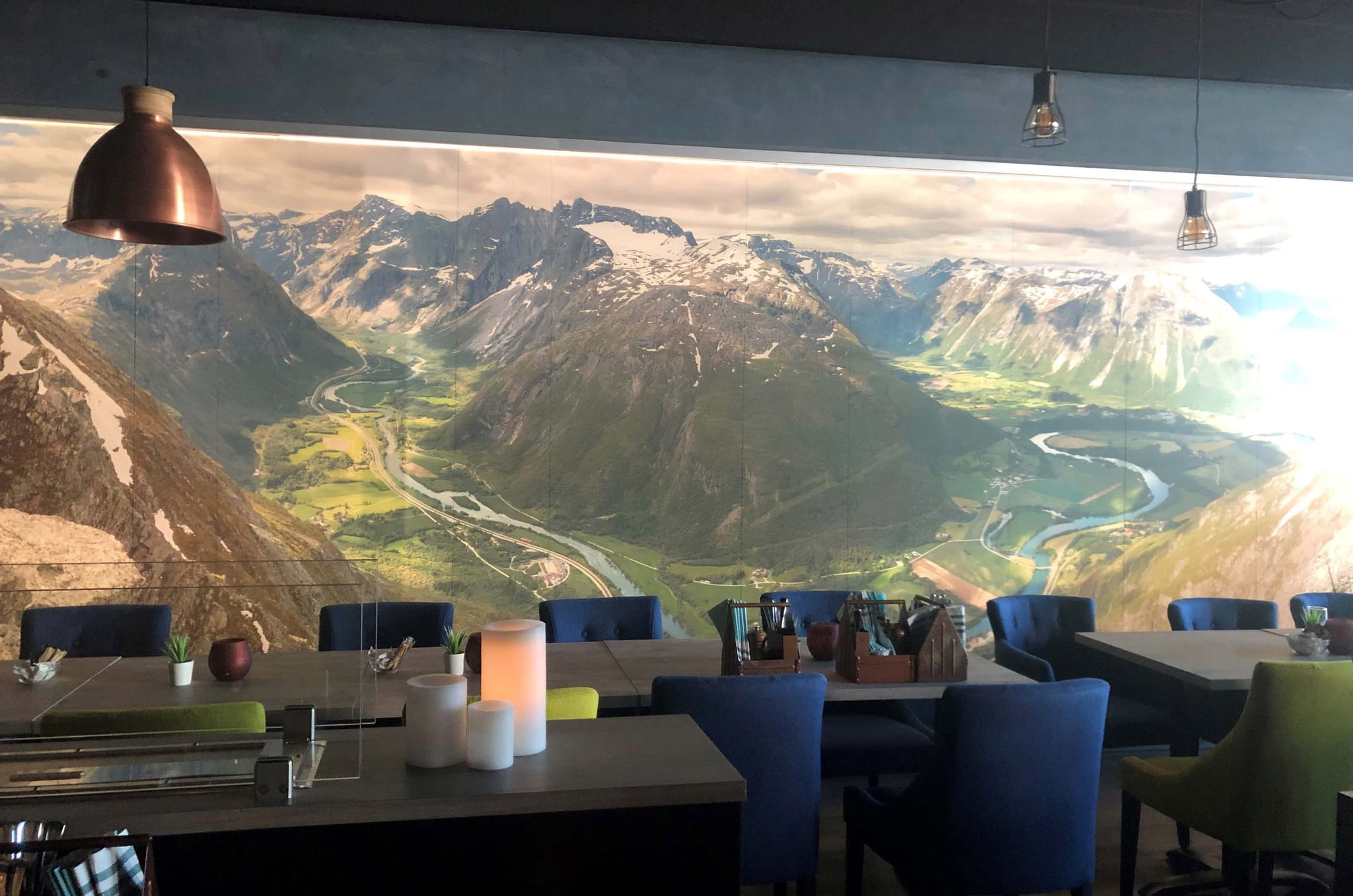 Long tables with blue and green chairs in a dark space with a long wall piece on the wall that is a picture of mountains and nature and has a lighting in the picture that makes the picture pop.