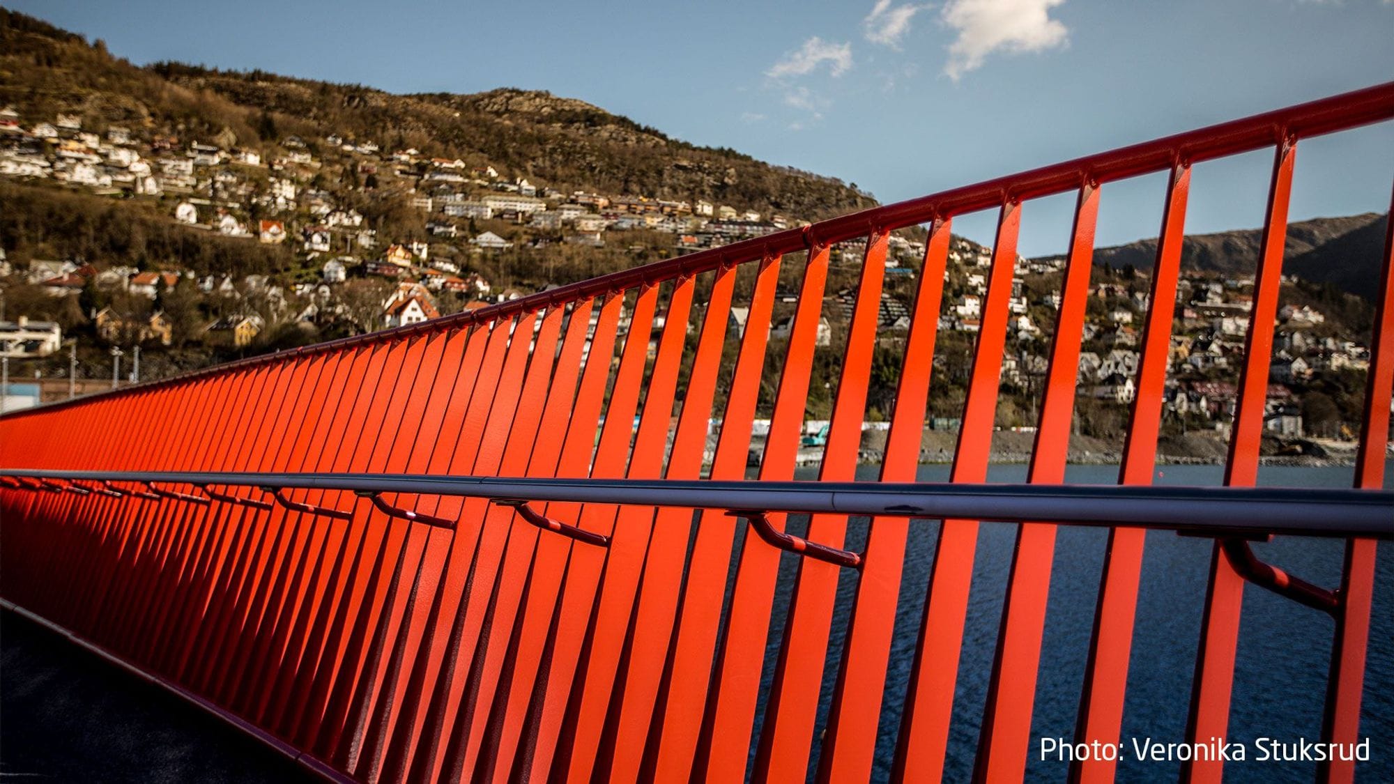 Closeup of a red railing on a bridge with a hill with houses in the background