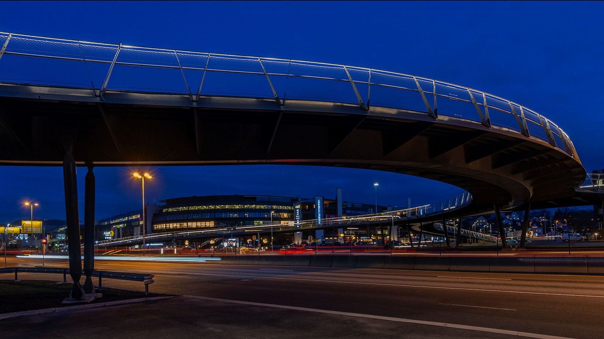 Under an arched footbridge and a street that goes under the bridge. In the background, the illuminated urban environment can be seen in the dark.