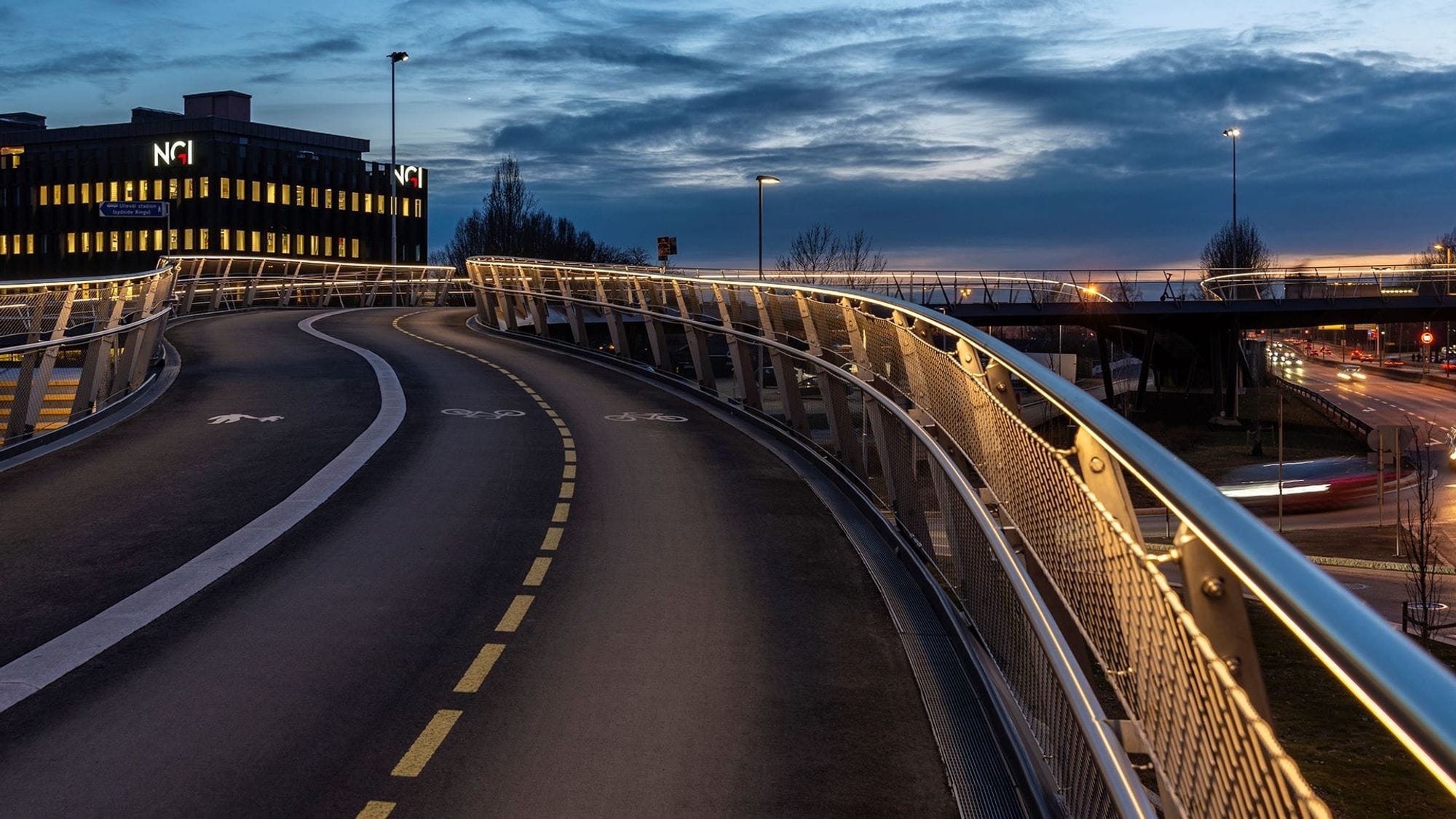 On top of a footbridge with three lanes in the dark. Street lights and lighting on the railing of the bridge illuminate the surroundings.