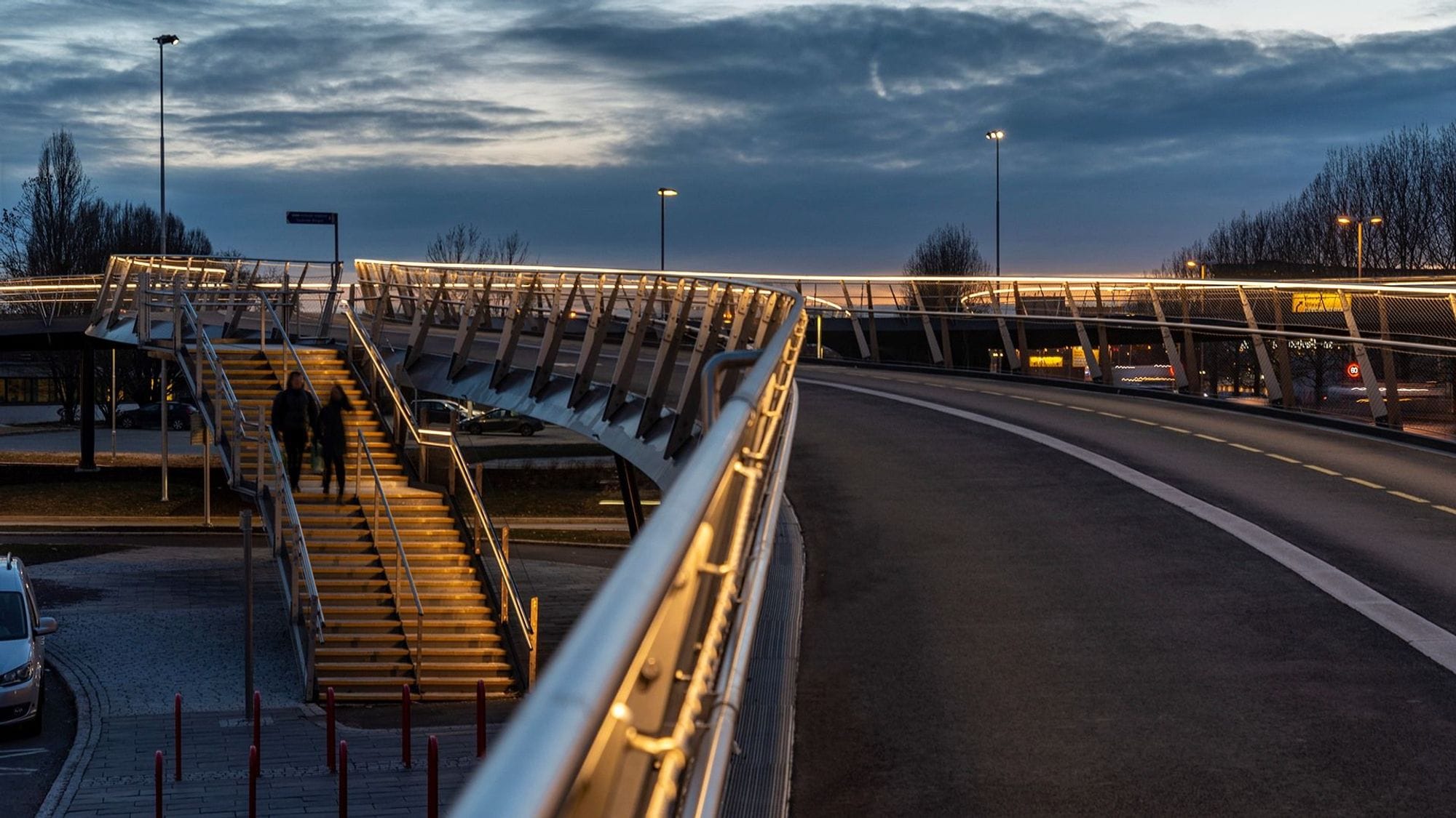 A bridge in the dark illuminated by lighting on a railing. Stairs down from the bridge with two people walking down.