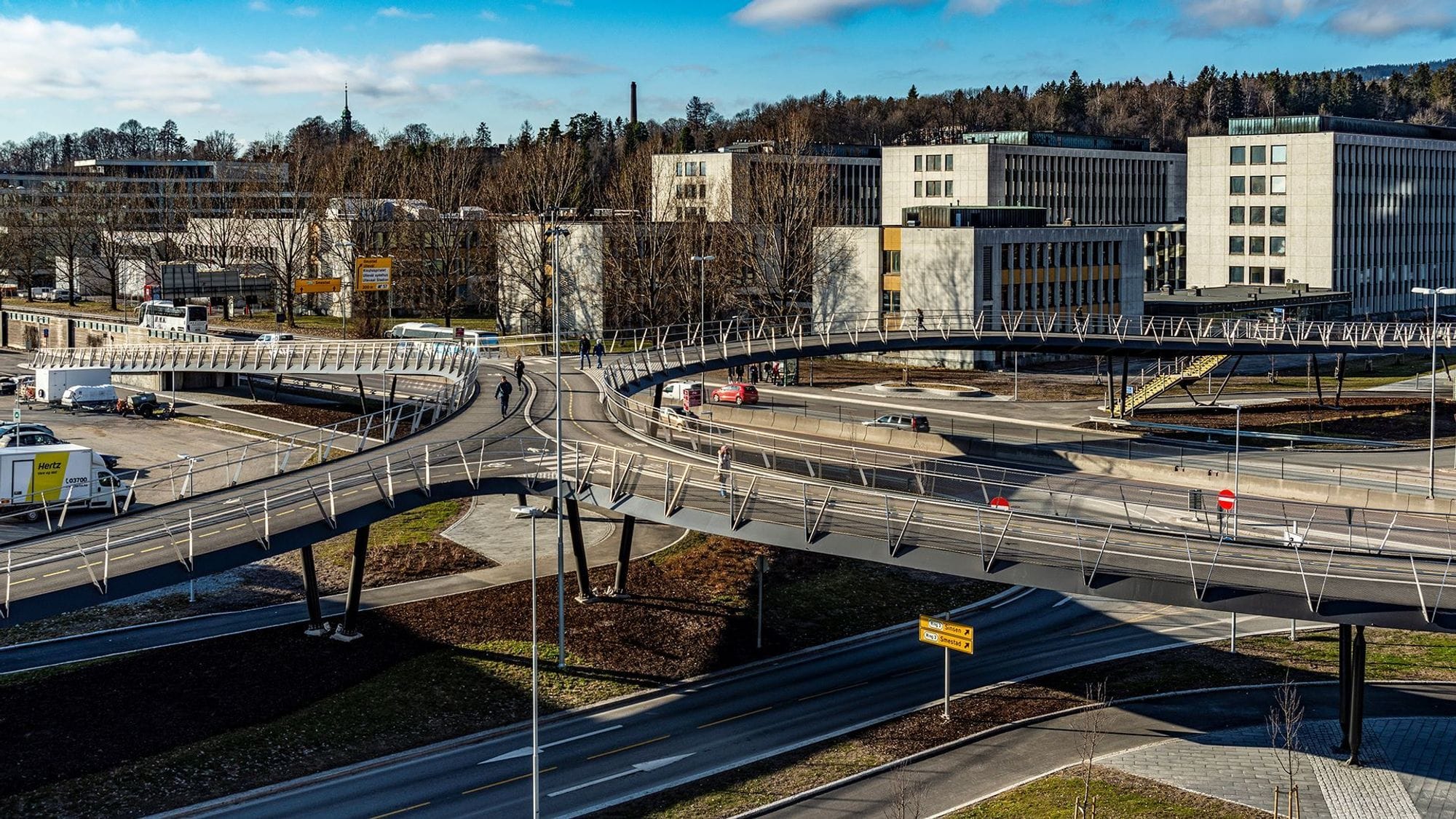 Footbridges meet over roads in a soft cross-shaped shape, and apartment buildings and vegetation can be seen in the background