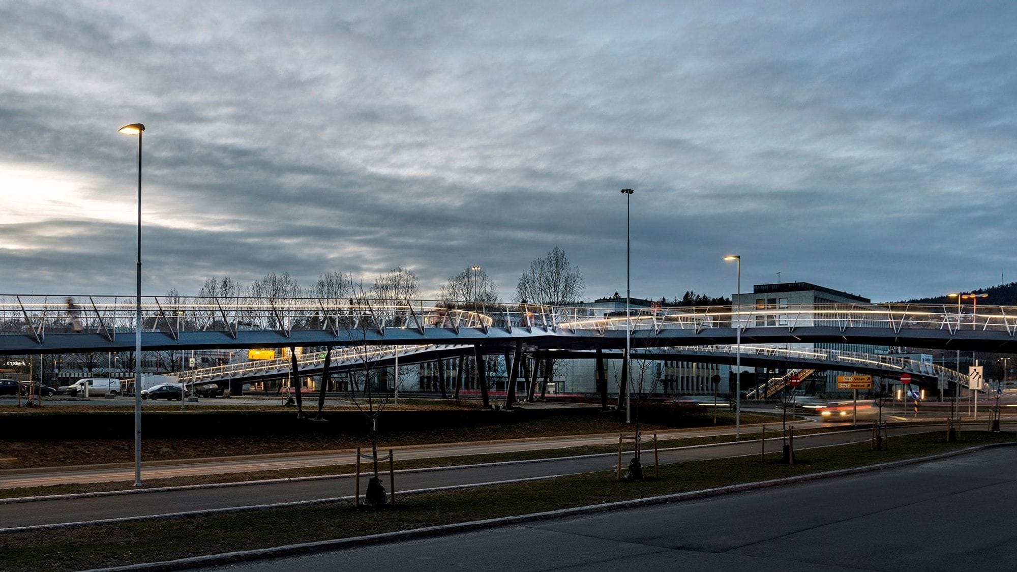Two lanes of the pedestrian and bicycle bridge meet over the road. Dark clouds lie over and the railing of the bridge is illuminated