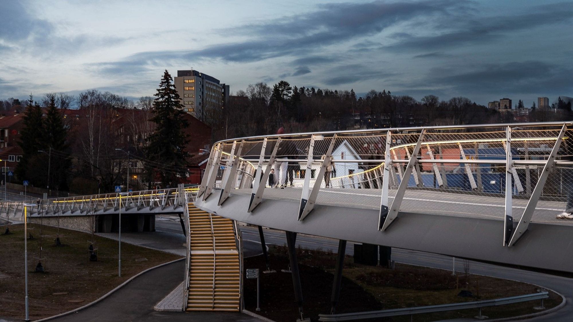 Close-up of the railing of a footbridge where the description of the railing is clearly visible and v-shaped structures on the railing. Steps up to the bridge can be seen in the background and people walking on the bridge