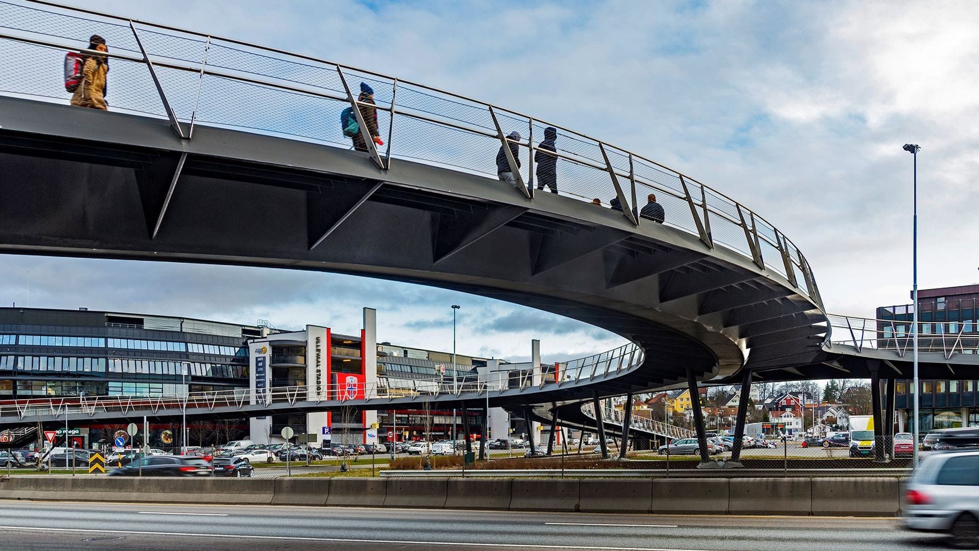 The pedestrian bridge can be seen under it with pedestrians using the bridge over a street with car traffic. In the background you can see buildings, cars and shops.