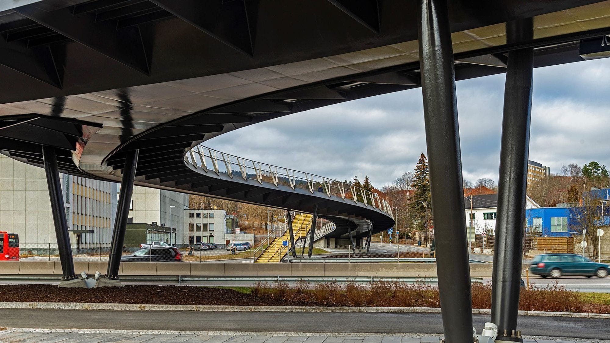 Under a footbridge by a street where cars drive under the bridge and houses can be seen in the background.