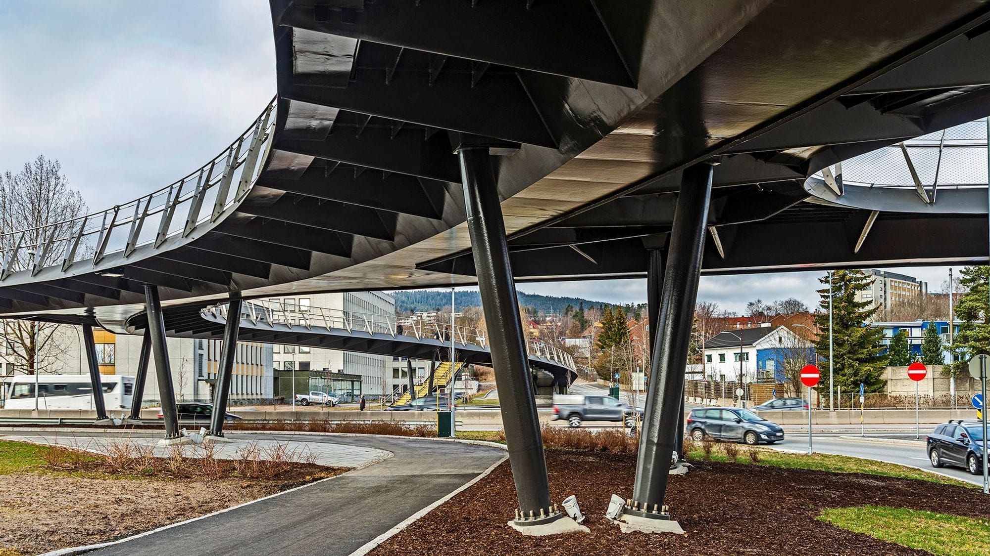 Picture showing under a footbridge with two load-bearing posts, a sidewalk on the left side surrounded by well-organized surfaces with grass, caves and tree beds. Cars drive on the streets in the background.