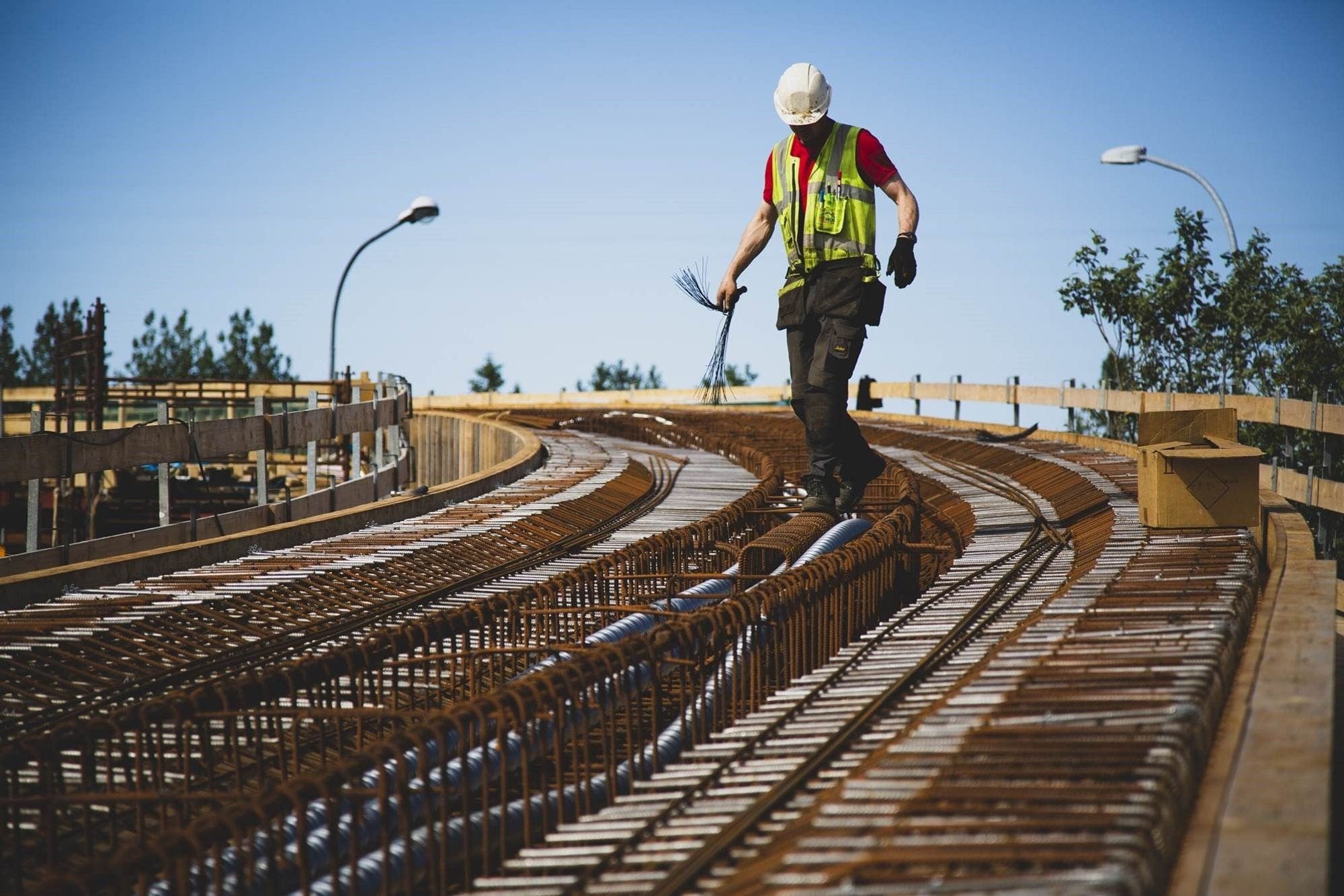 A man wearing black paints, red t-shirt under a yellow working vest and has a white security helmet on working on a bridge ynder construction