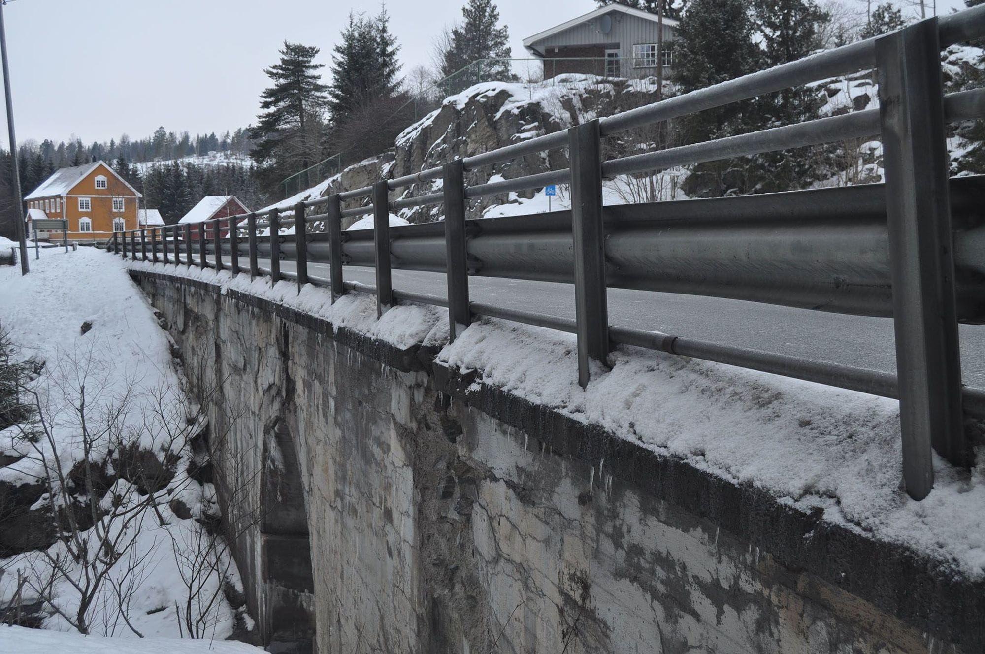 Berger bridge before rehabiliation. Close up of Retaining concrete wall with old railing on top of the bridge in snowy surrounding with trees and two houses in the background