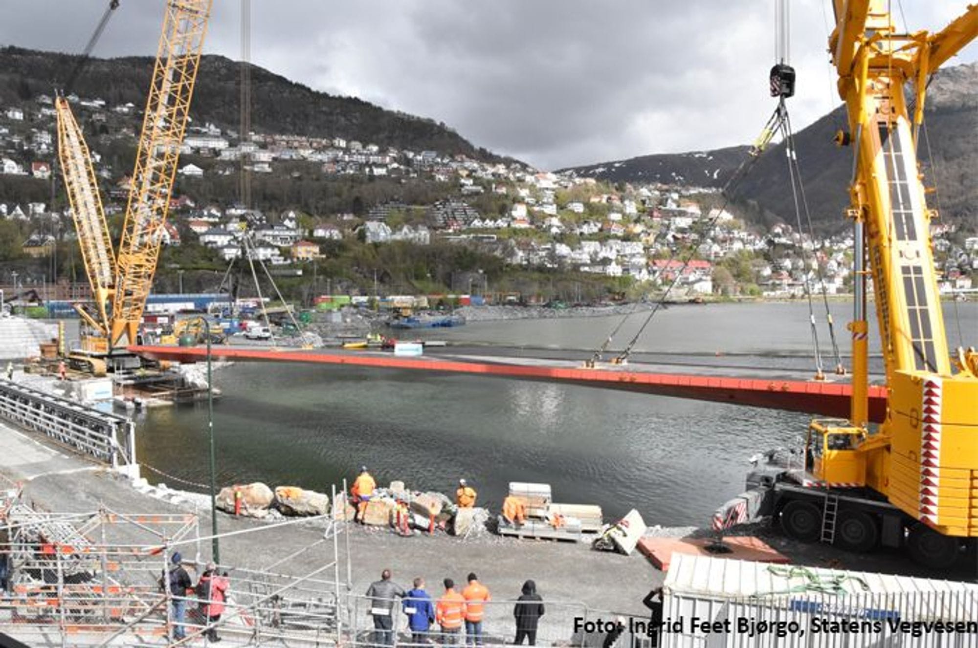 Men in work clothes watch two yellow cranes lift up a red bridge to locate it over the sea