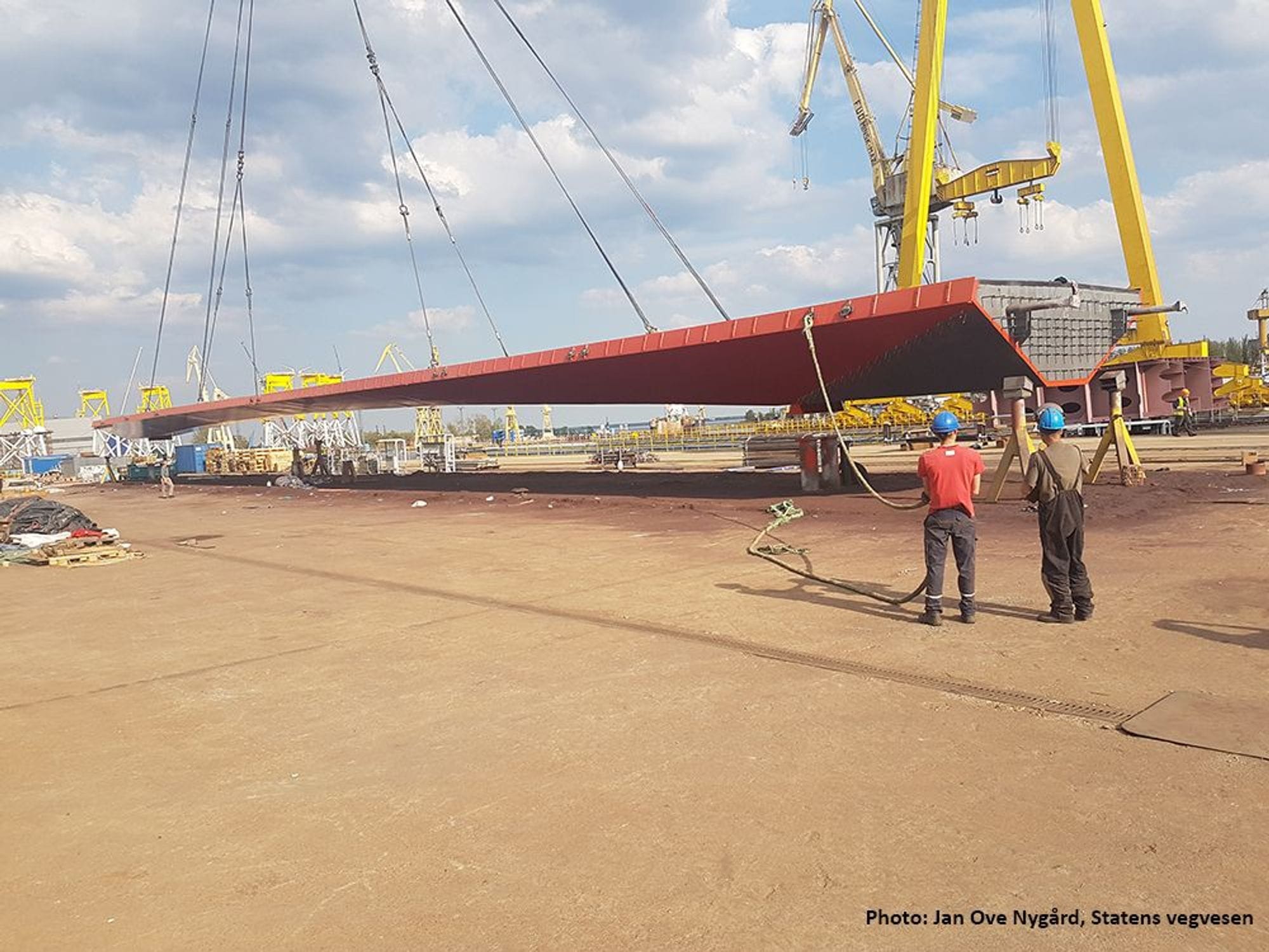 Two workers watch a crane lift up a large and red steel bridge on a construction site