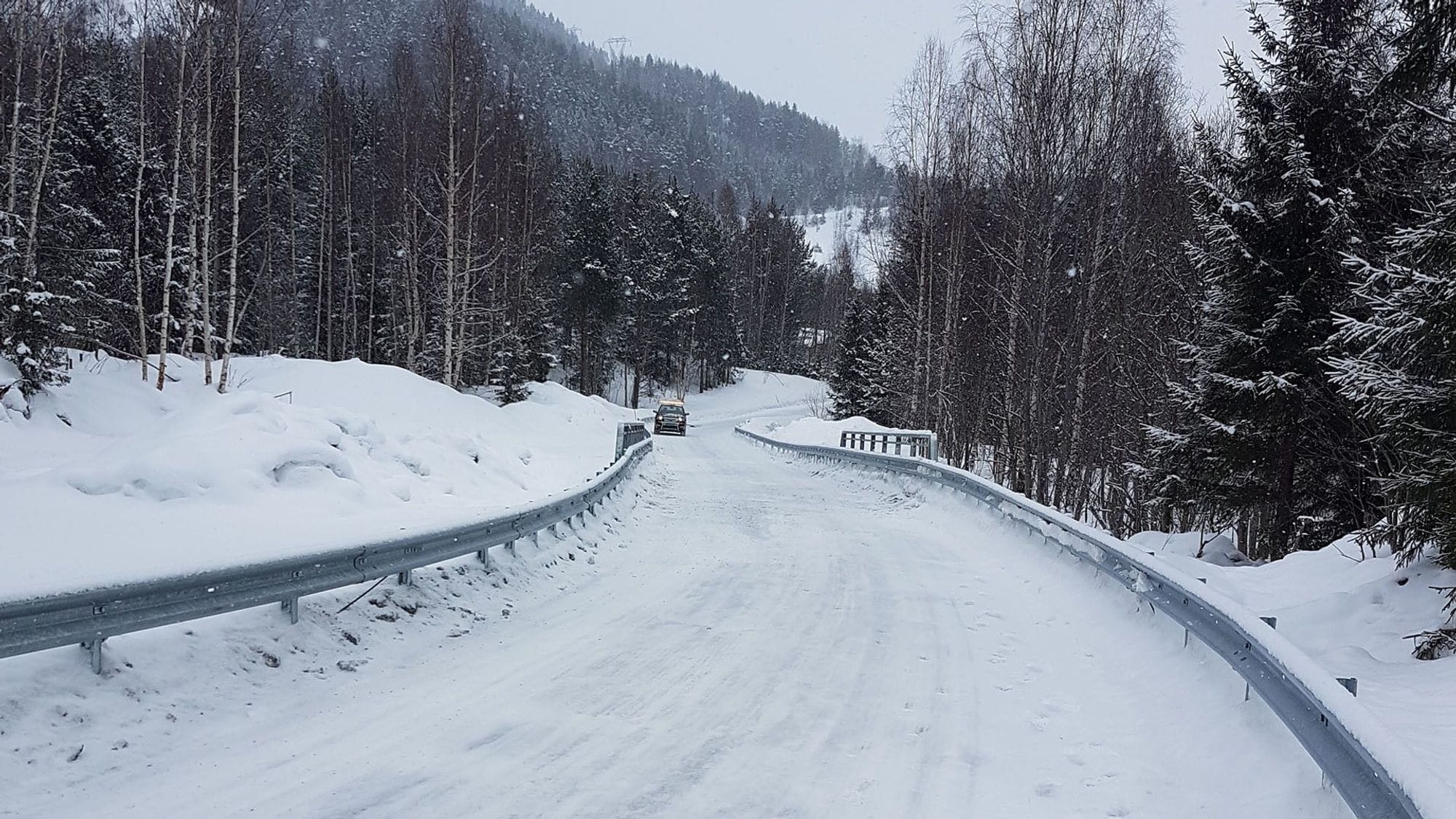 Snowy road seen over Velta bridge surrounded by forest. New railings and wider guide to the bridge.
