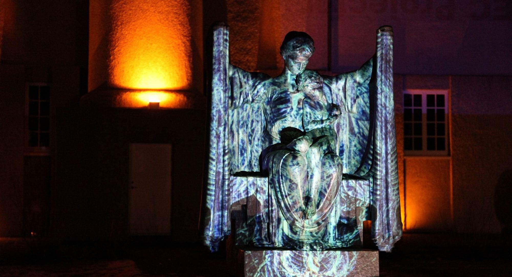 A statue of a woman holding hands up and has a child in her lap. The statue has purple, blue and green lighting casted on it.