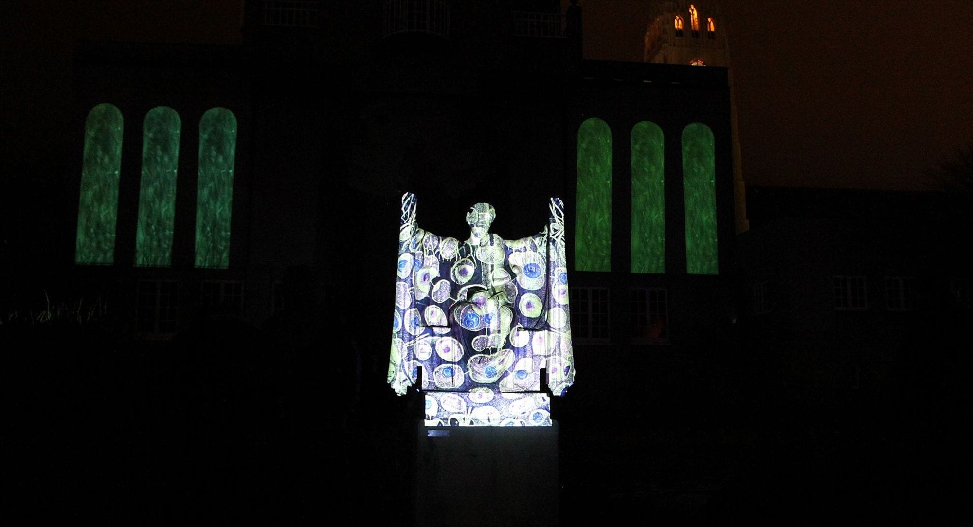 A statue of a woman holding hands up and has a child in her lap. The statue has purple, blue and green lighting casted on it.