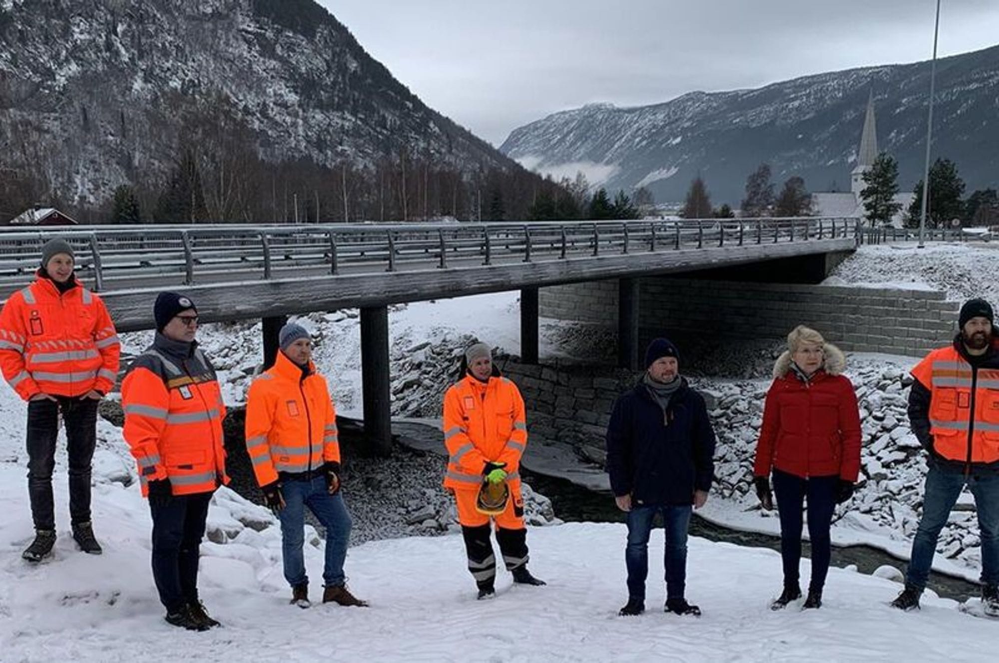 People standing in front of the new bridge, five in orange work jackets and two persons in black and red jackets.