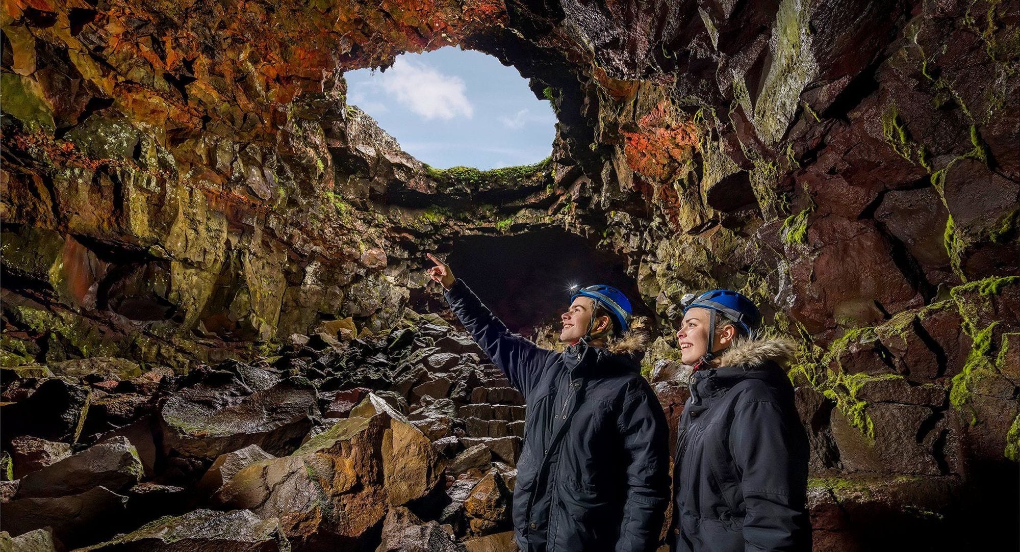two people with security helmets standing in a cave looking and pointing towards the cave wall and a hole in the cave shows blue sky