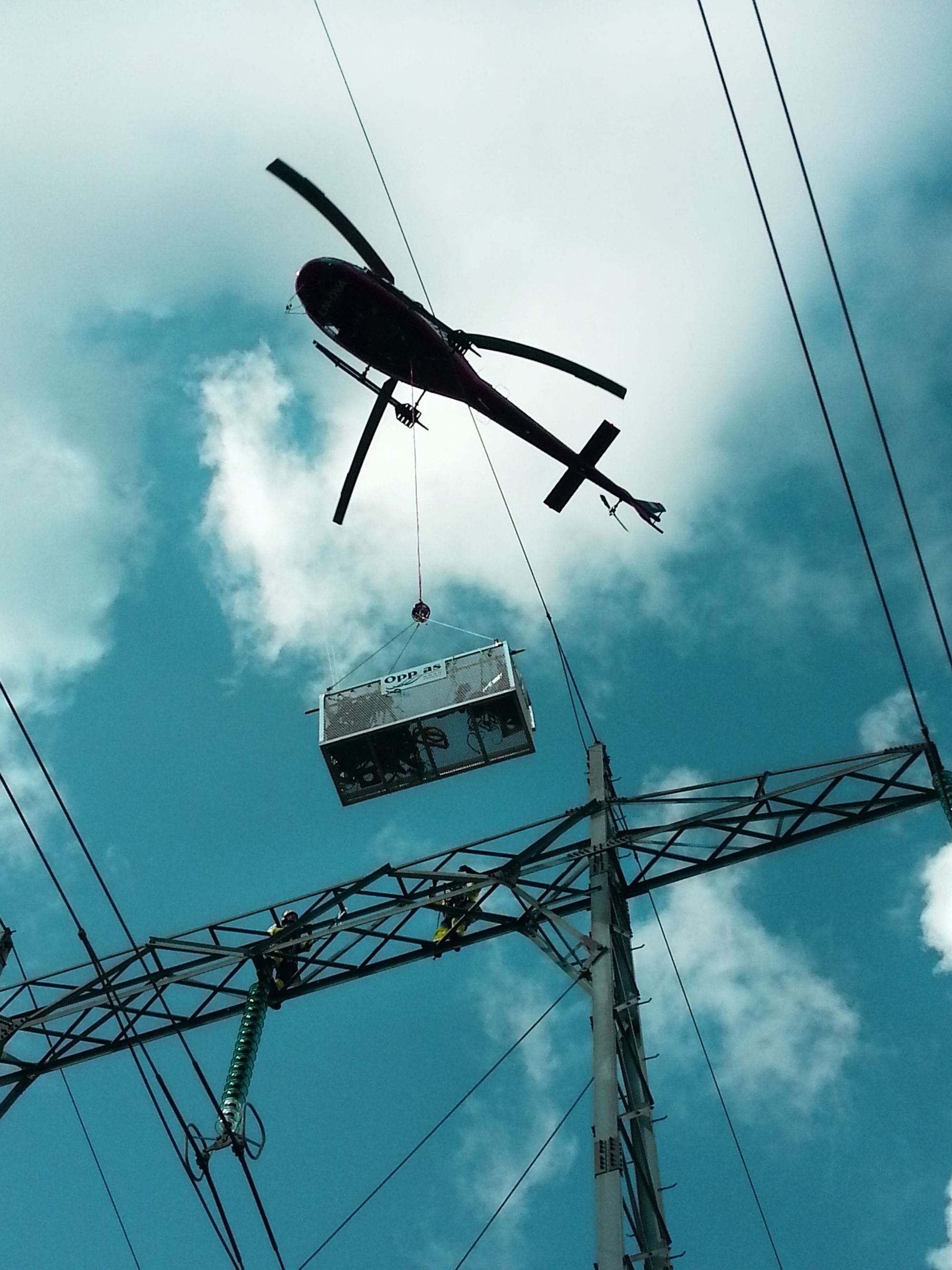 Helicopter flying with a box over an electrical mast, blue sky and clouds above
