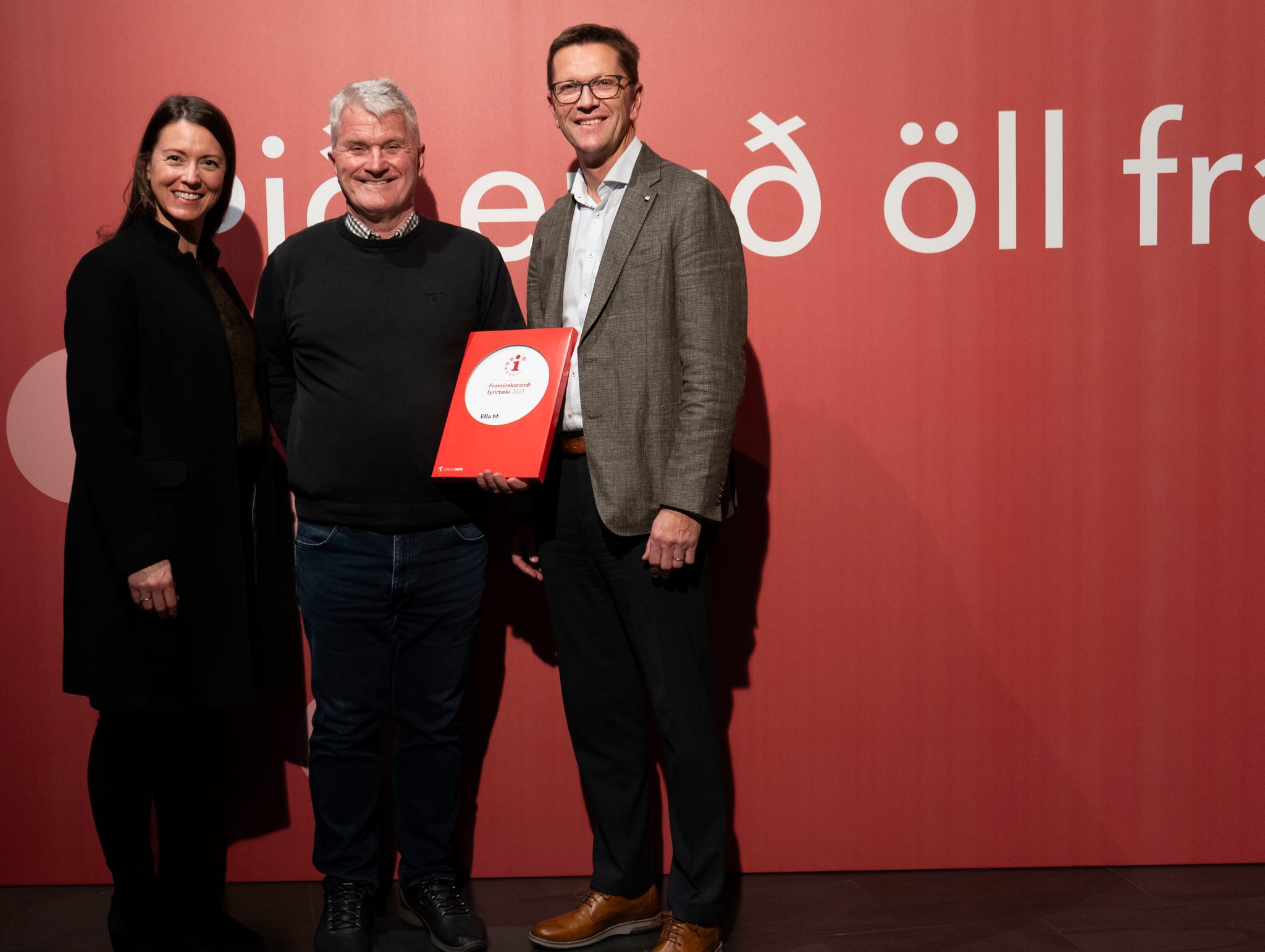 A woman and two man standing in front of a red background holding a red award certificate and smiling