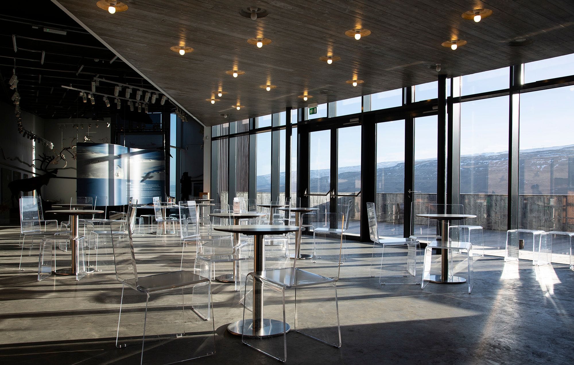Inside a banquet hall, windows on one side, lights hanging from the wood-clad ceiling, round tables and easy chairs. Scandinavian look
