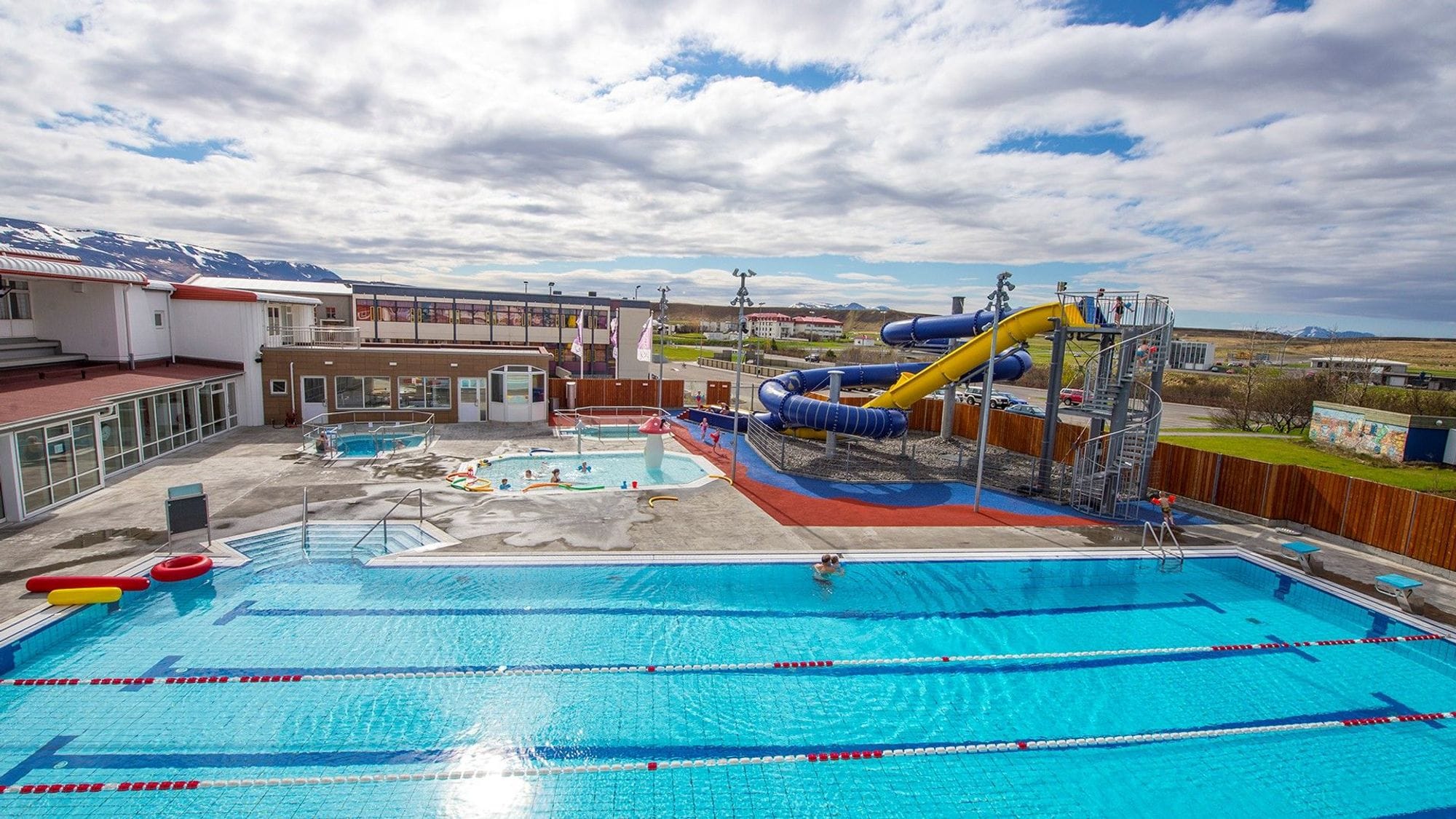 View of a pool garden with cloudy skies. One large pool, two large slides, one blue and the other yellow, along with two tubs and one shallow children's pool with colorful pool toys. A pool building surrounds the corner of the pool garden.