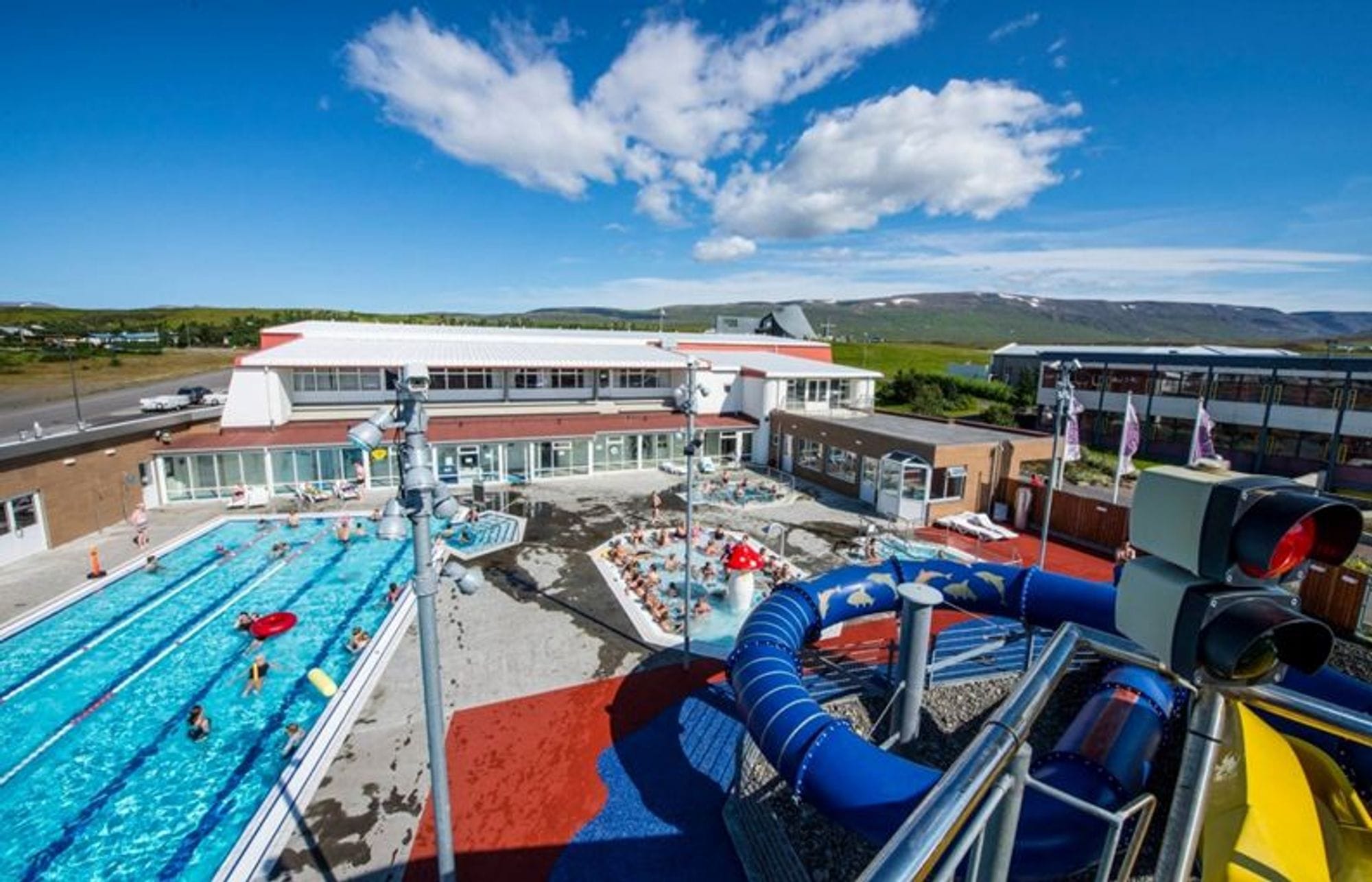 Overview of a pool area with one pool with people plus a hot tub full of people. Large blue slide and paved plot. A pool building seen in the background along with a blue sky with just a few clouds.