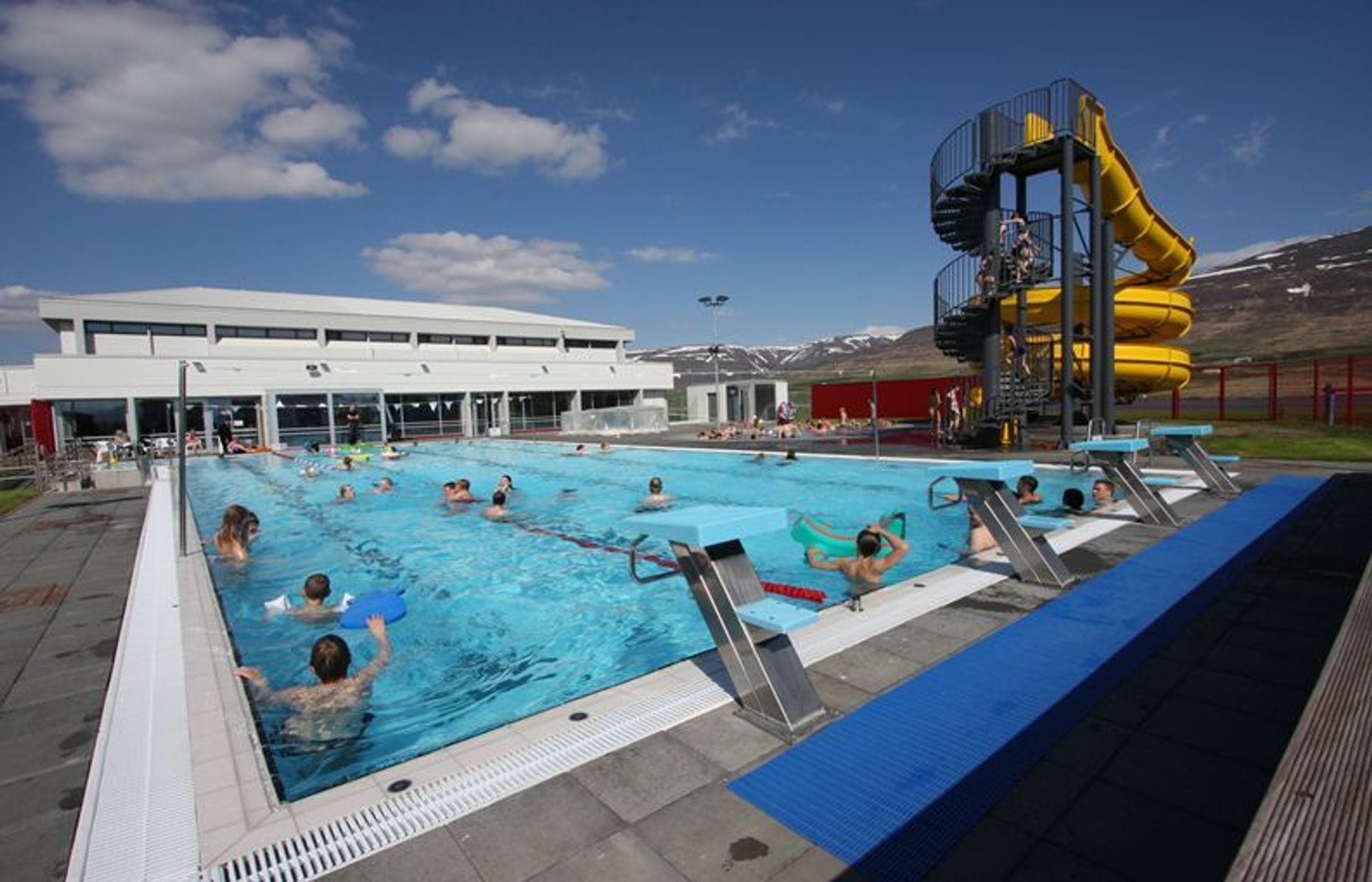 One large swimming pool with jumping platforms. People are in the pool. Large yellow slide with a circular staircase to the side. White pool building and blue sky.
