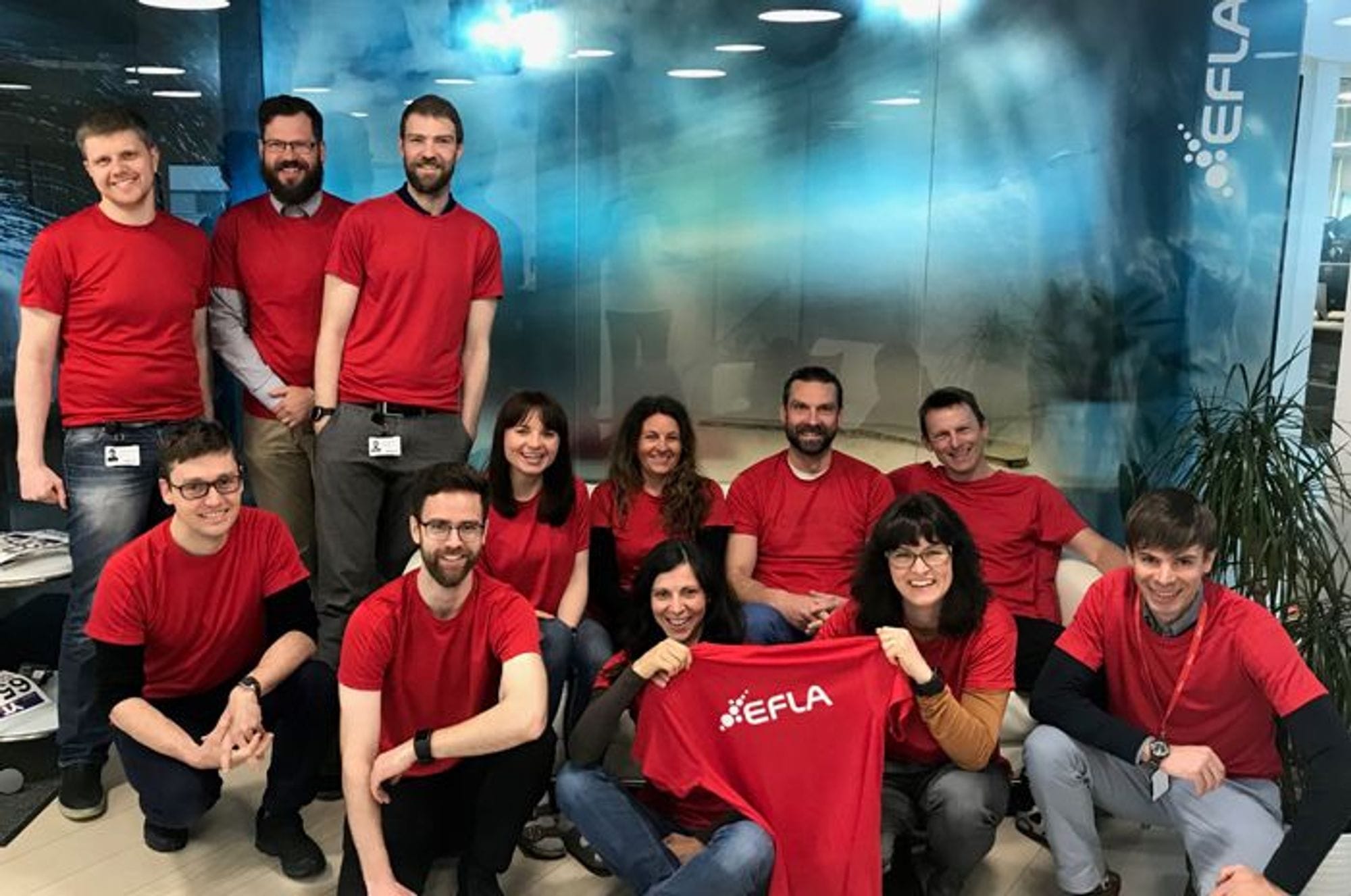 People wearing red t-shirts sitting and standing in a group and two of them holding a red t-shirt showing EFLA brand on the shirt