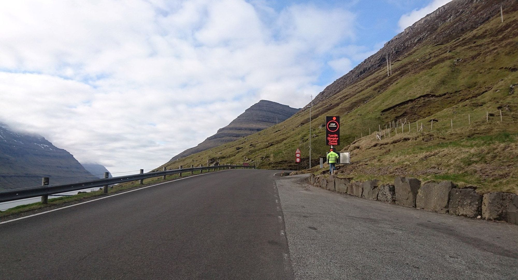 A road leading to a tunnel that leads through a mountain, a man in a yellow work jacket stands by a black and red sign next to the entrance to the tunnel