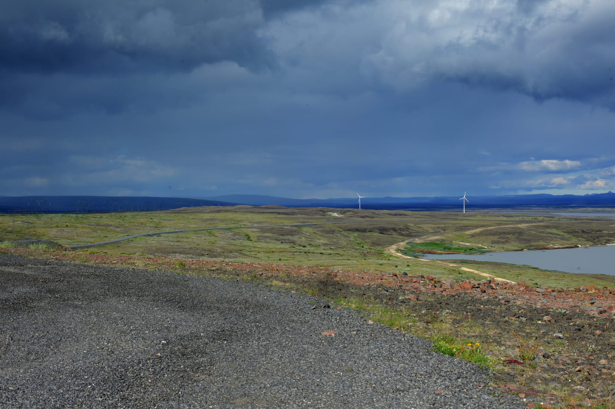 Looking over a landscape, gravel in the foreground and then grassy fields with water on the left side. In the distance you can see two windmills and behind them blue mountains