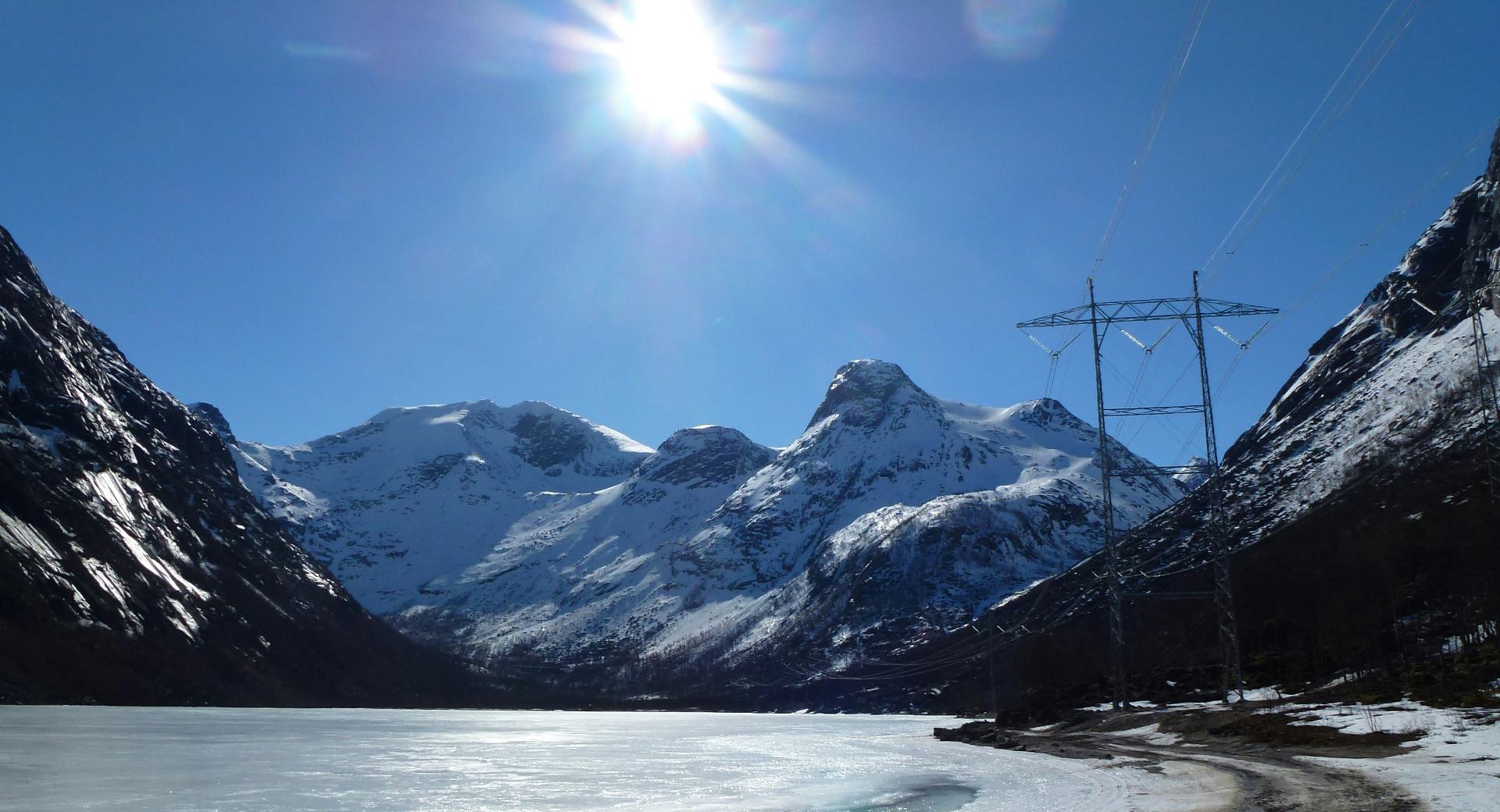 Bright sun reflecting on snow covered mountain, lake and transmission tower