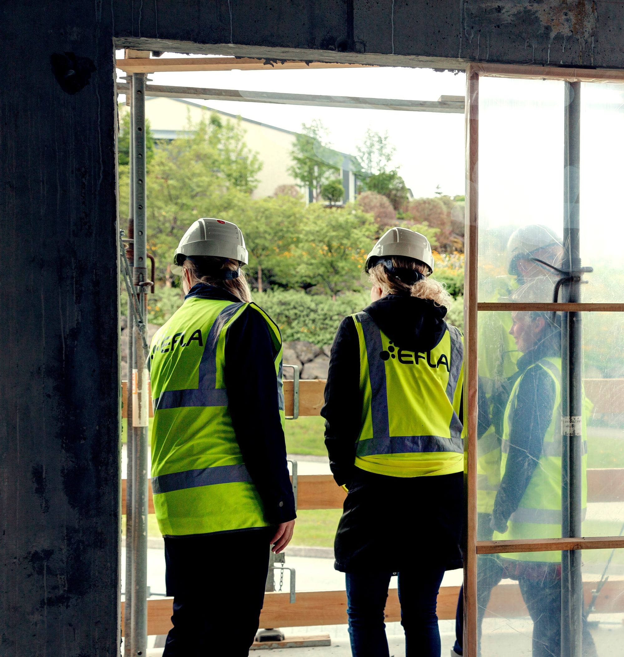 Women in yellow safety wests with helmets looking outside a window that's in construction. 