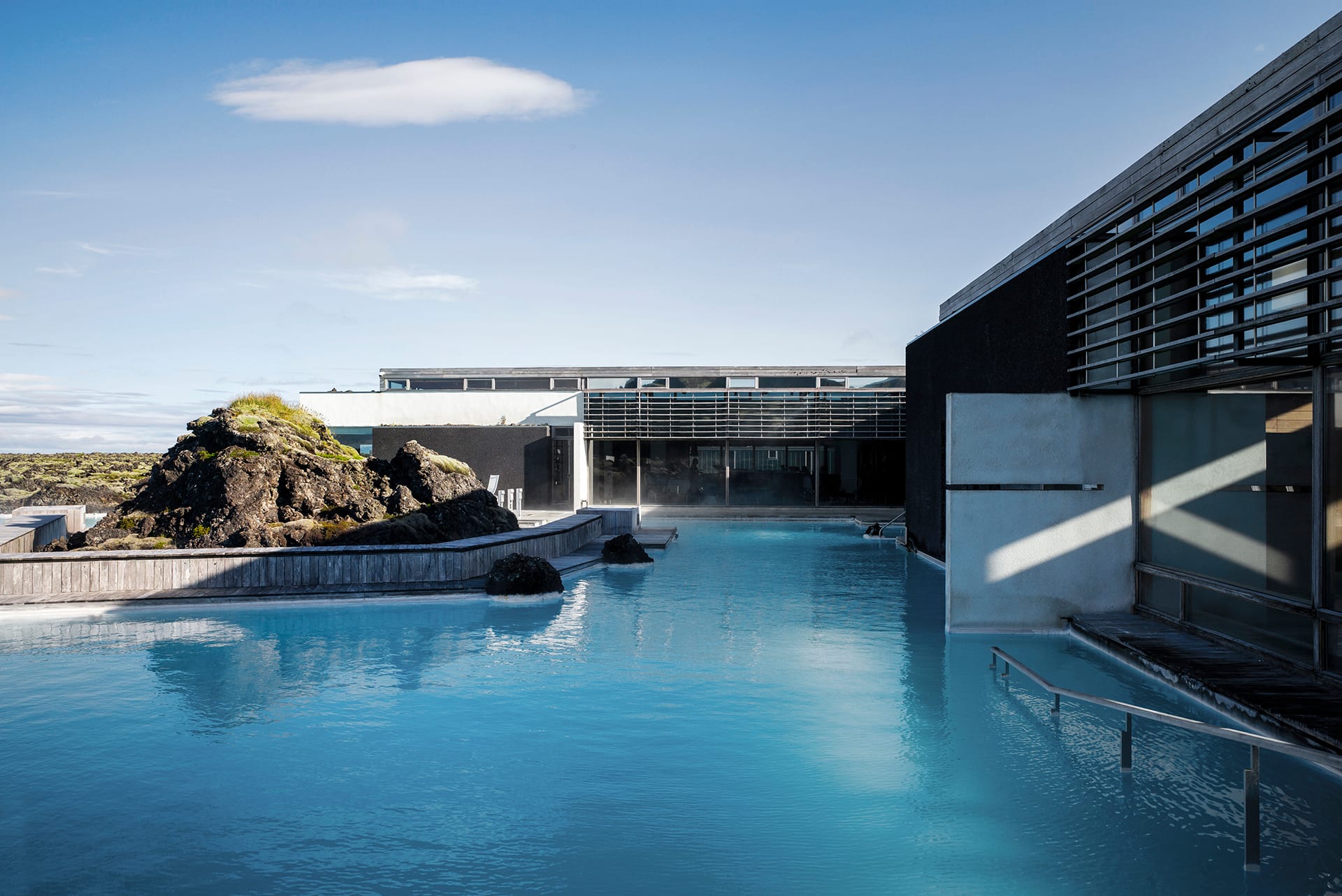 A blue lagoon in the foreground, an L-shaped concrete and wooden building on the right and in the center of the picture at the back, lava and moss on the bank on the right