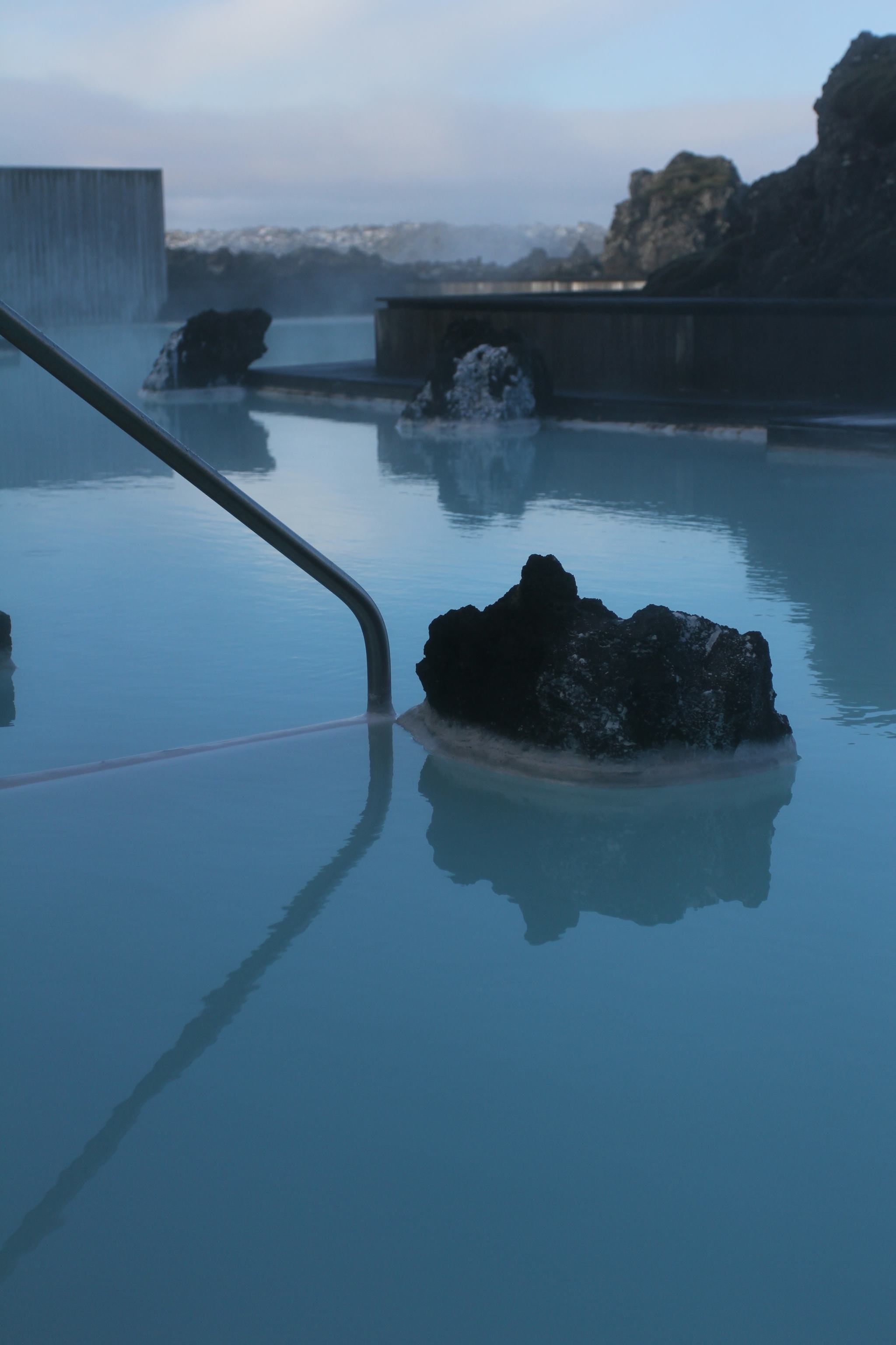 Blue lagoon, steam rises from it. Railing on the left side and lava lump in the middle above the lagoon