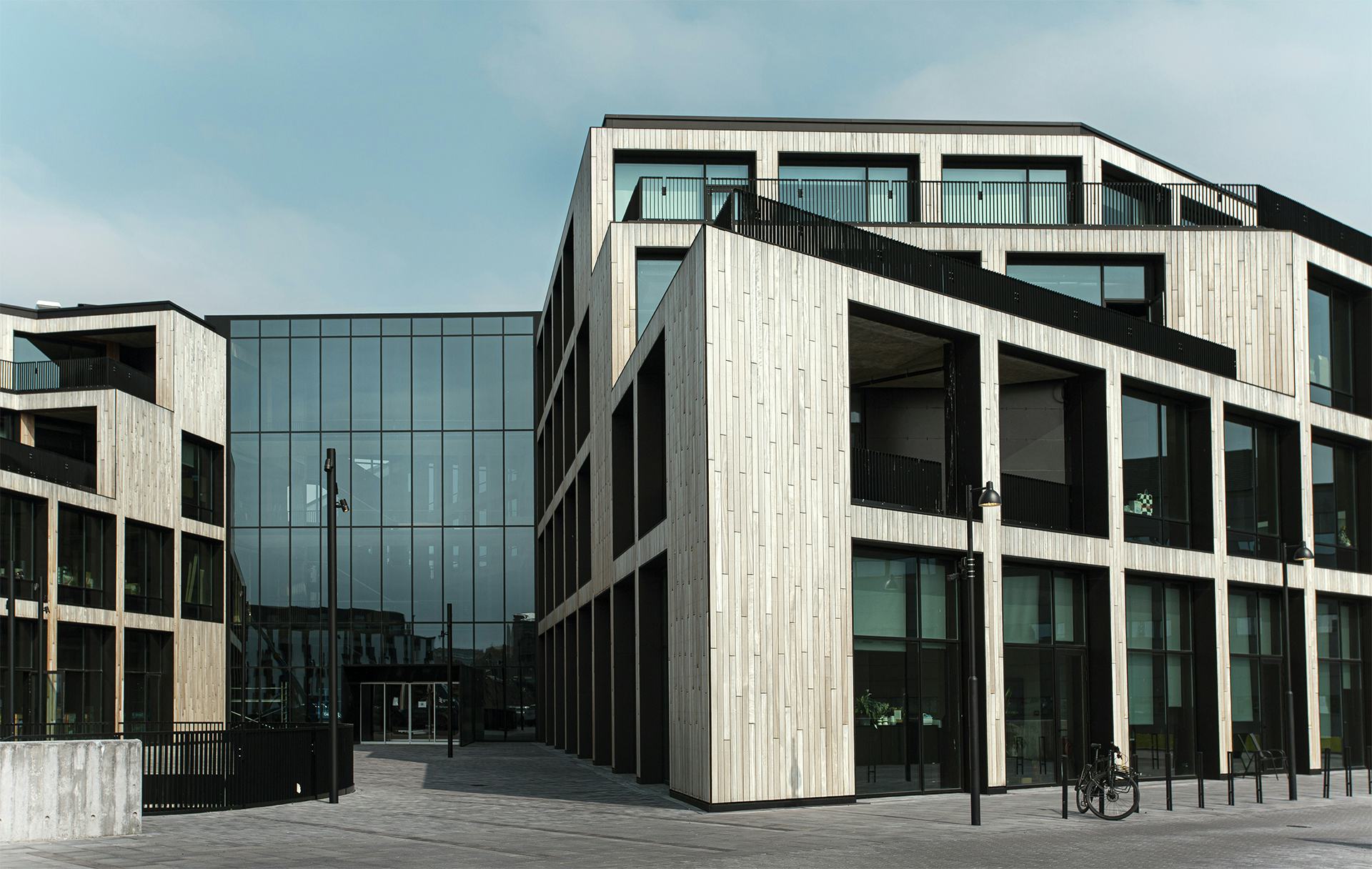 Looking at the front of a modern building with sharp corners. The building is made of wood and glass and stands on a paved square
