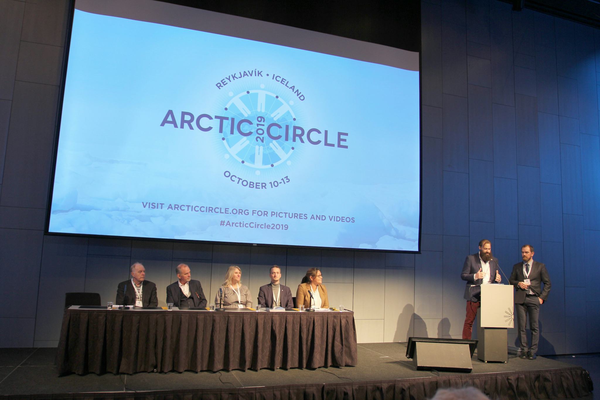two men delivering a speech at an Arctic circle conference, with five individuals seated behind them