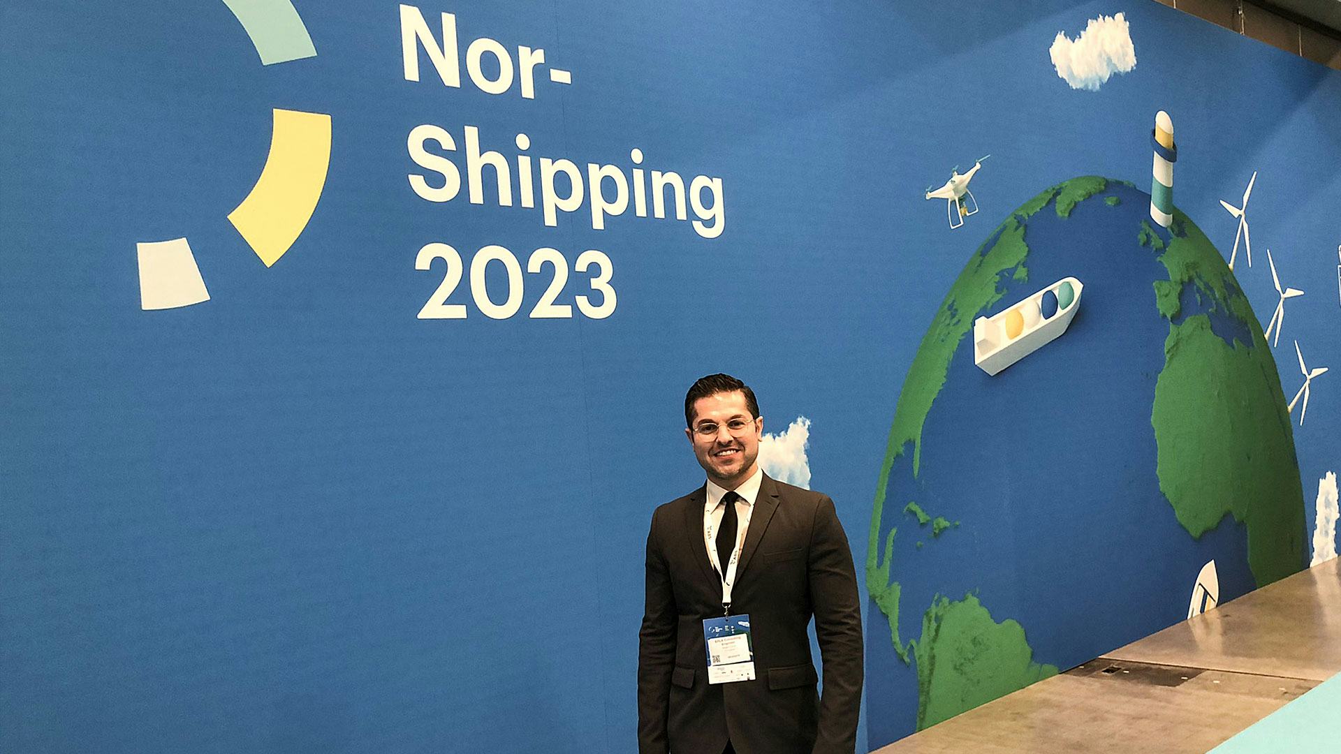A man in black suit standing in front of Nor-shipping 2023 poster wall